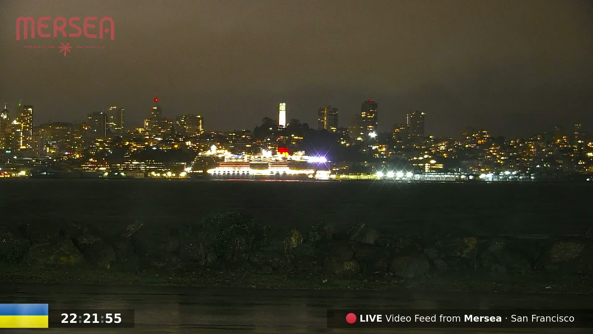 A nighttime view of a city skyline across a body of water, featuring a large cruise ship, under a dark, possibly overcast sky.