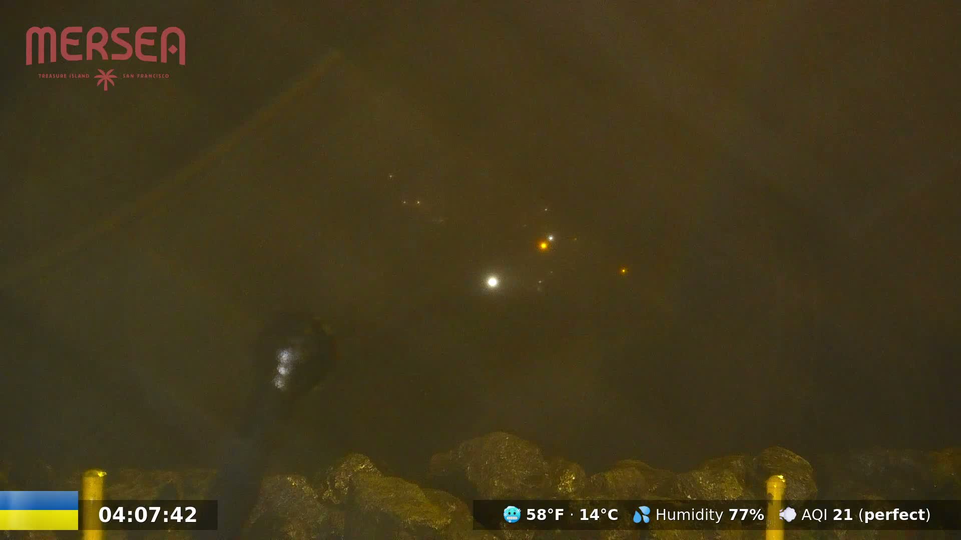 A dark, foggy nighttime scene shows a body of water with dark rocks in the foreground and a few faint lights visible in the distance.