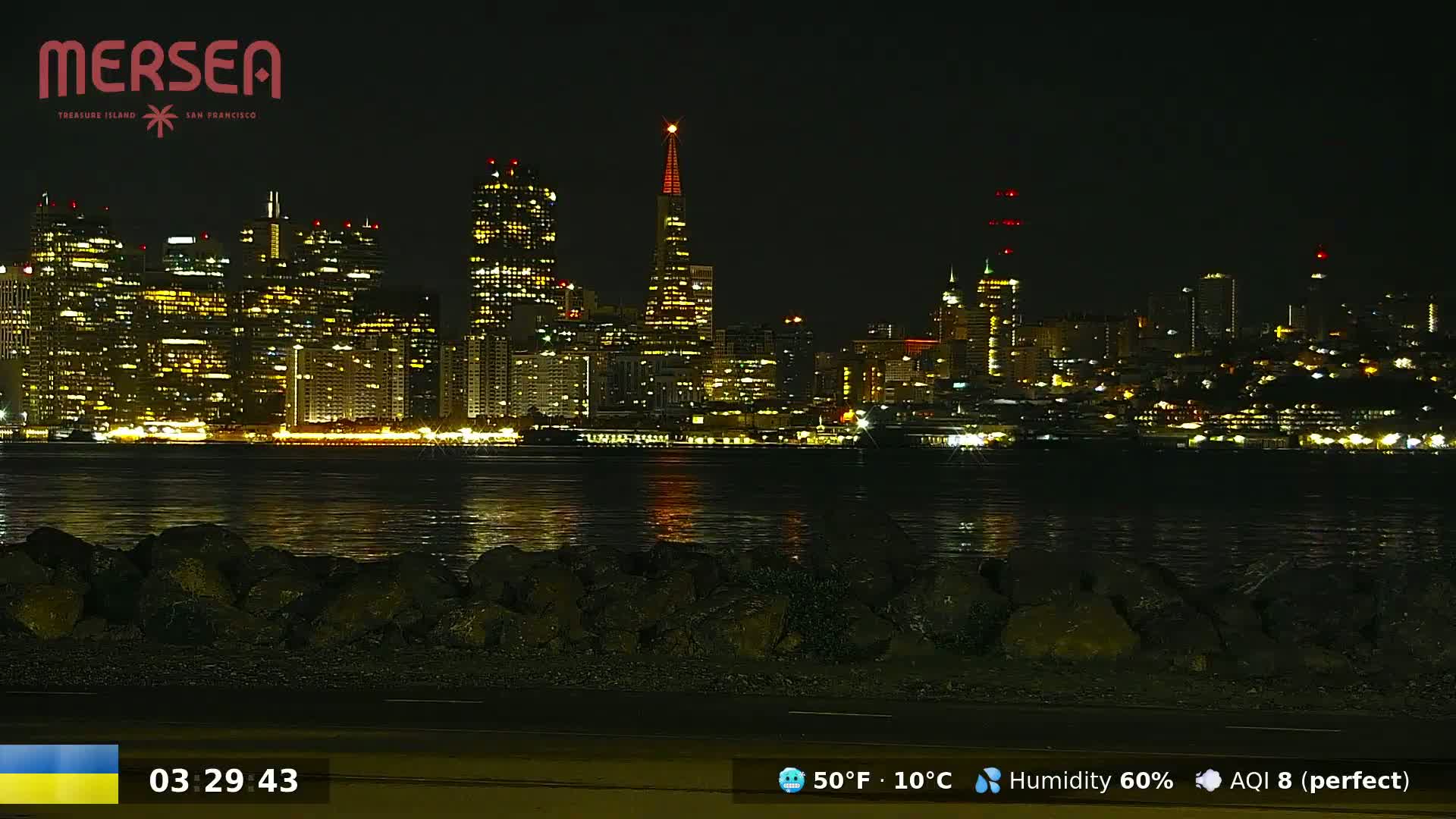 A nighttime view of a brightly lit city skyline across a calm body of water, viewed from a rocky shoreline.