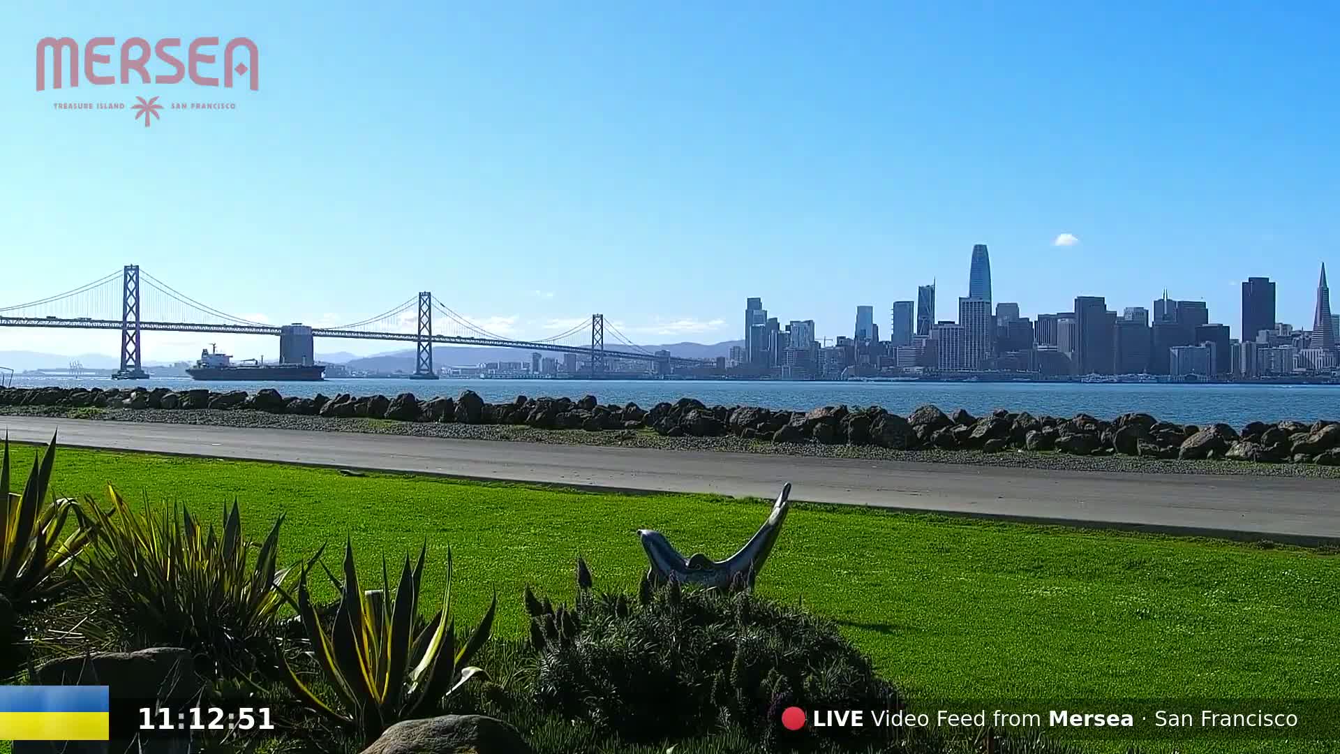 A grassy park with a sculpture overlooks a bay, a large bridge, and the San Francisco skyline under a clear blue sky.