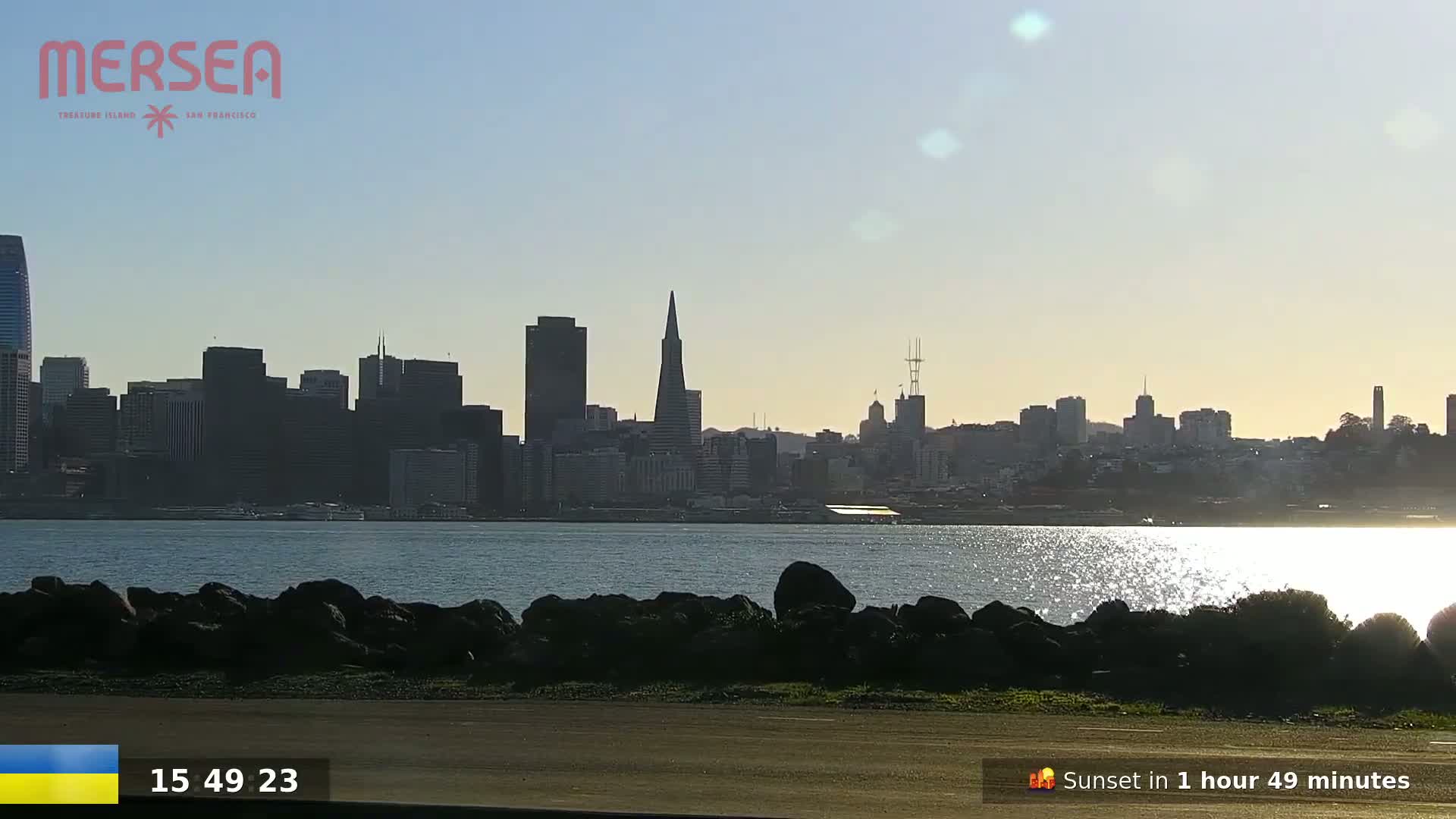 A sunlit city skyline is visible across a calm body of water from a rocky shoreline.