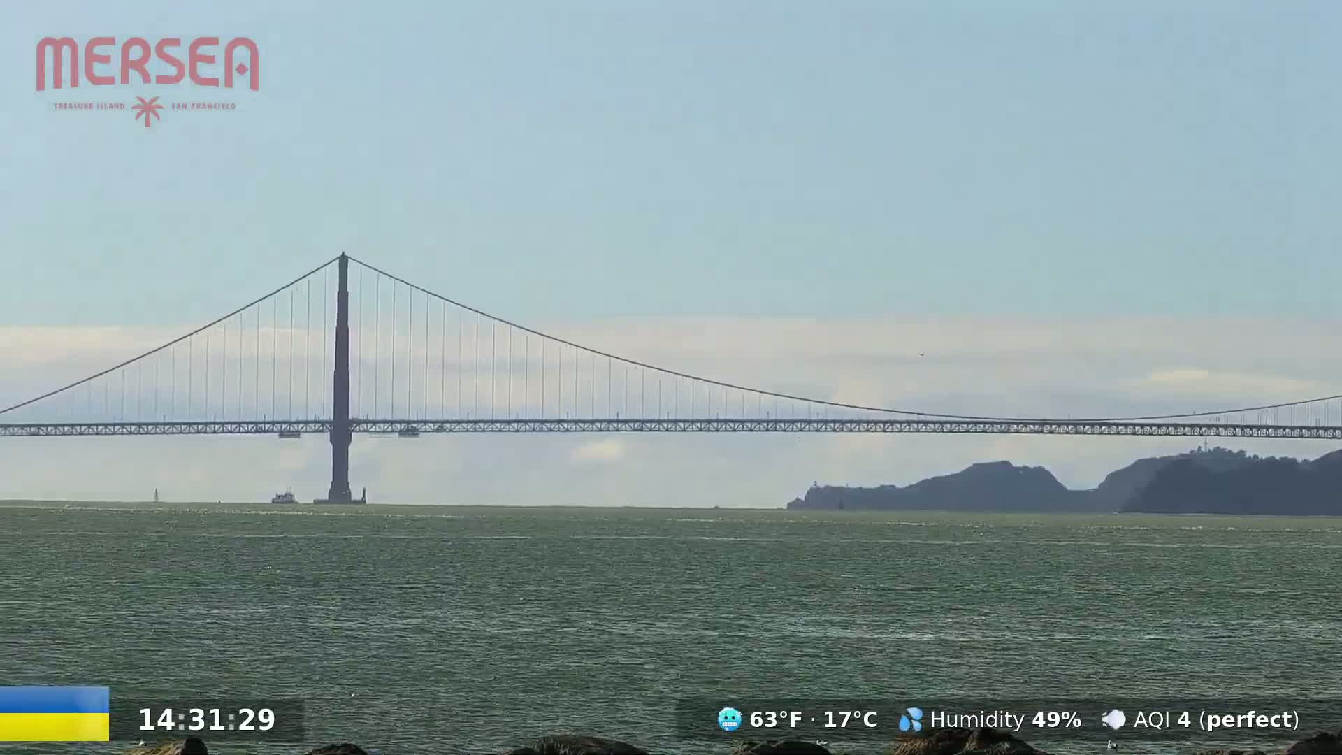 A suspension bridge spans a body of water under a mostly clear sky with a few faint clouds.