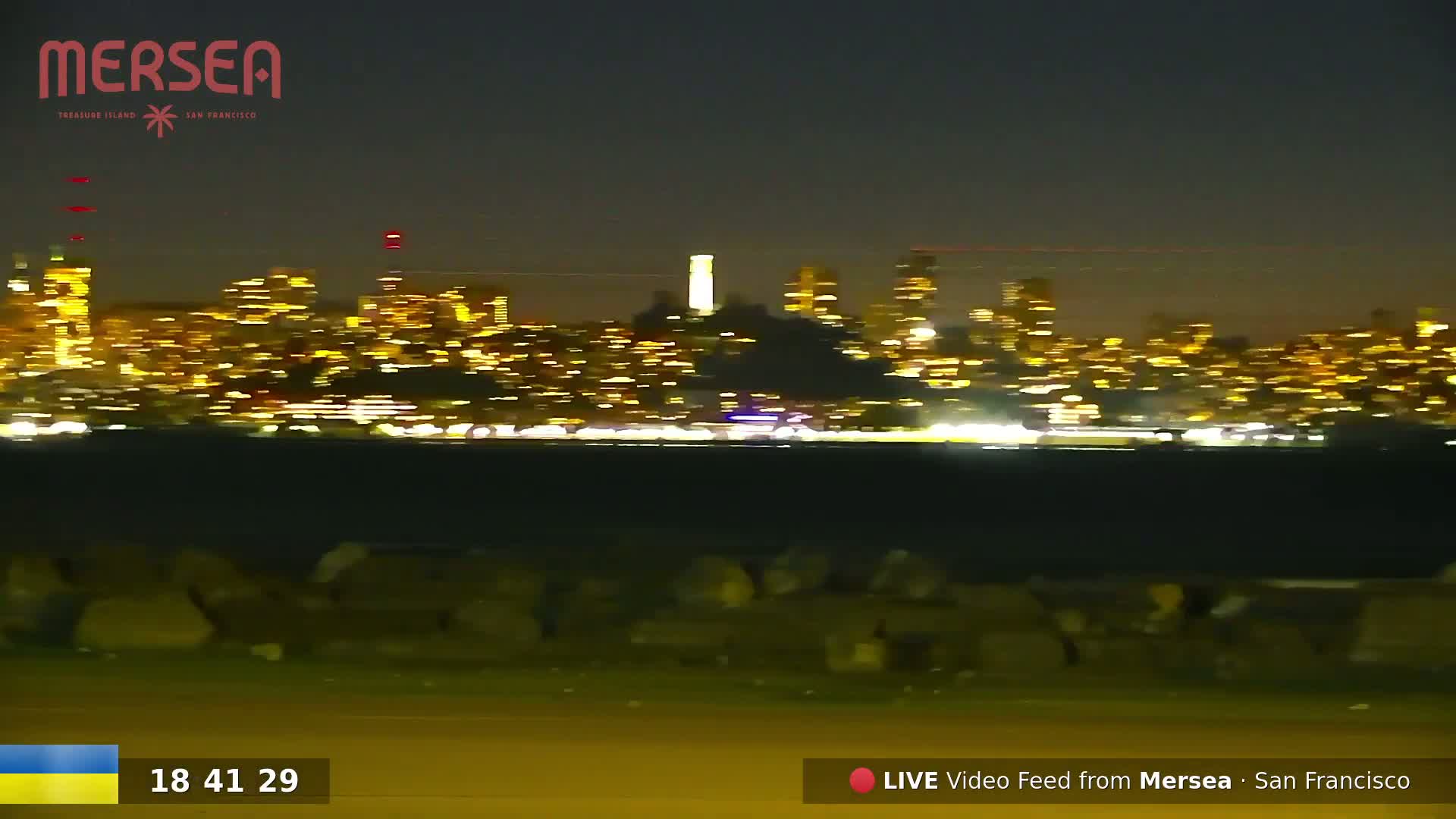 A nighttime view of a city skyline illuminated by numerous lights across a body of water, with a group of people gathered in the foreground.