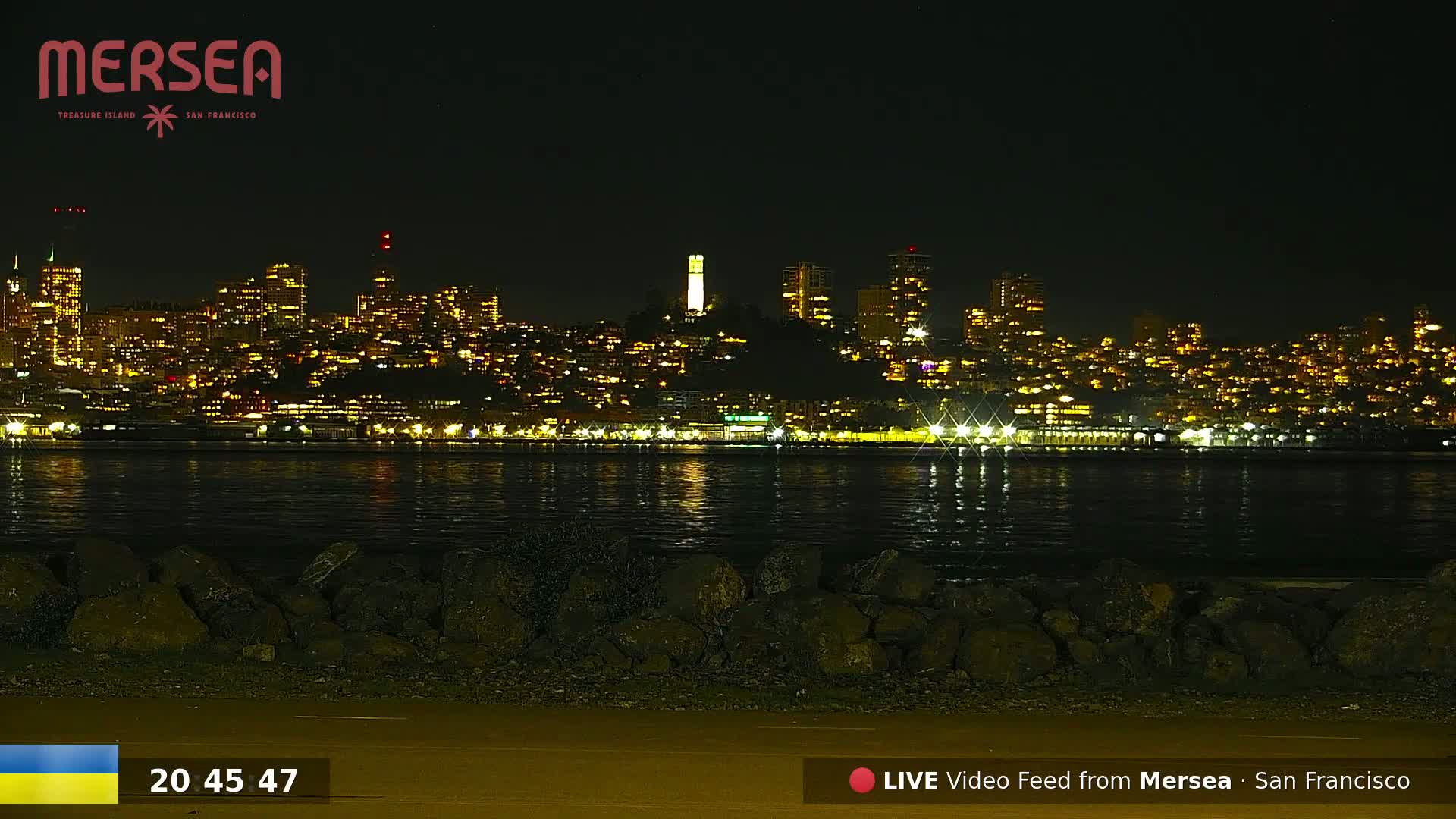 A nighttime view of a city skyline across a body of water, with the city lights reflected on the calm water and a rocky shoreline in the foreground.
