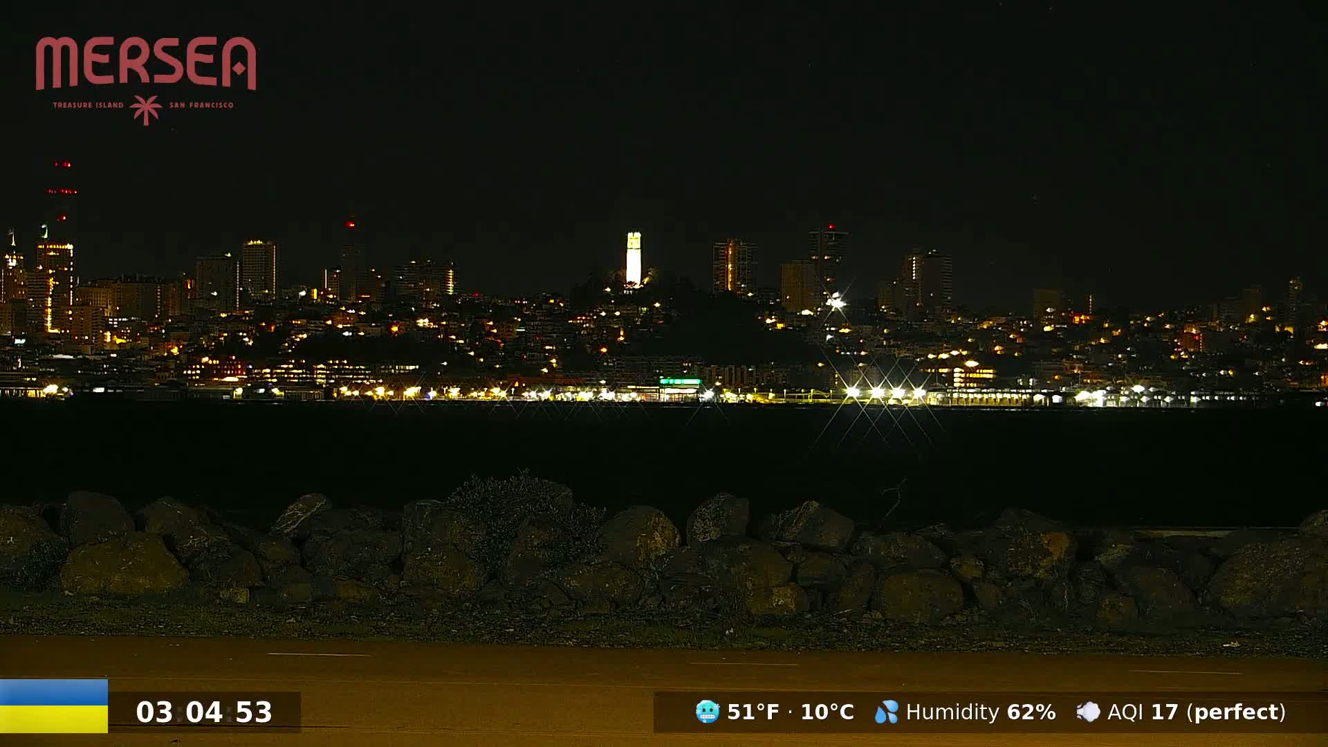 A nighttime view of a city skyline across a body of water, featuring a prominent tower, with a rocky foreground under a clear sky.