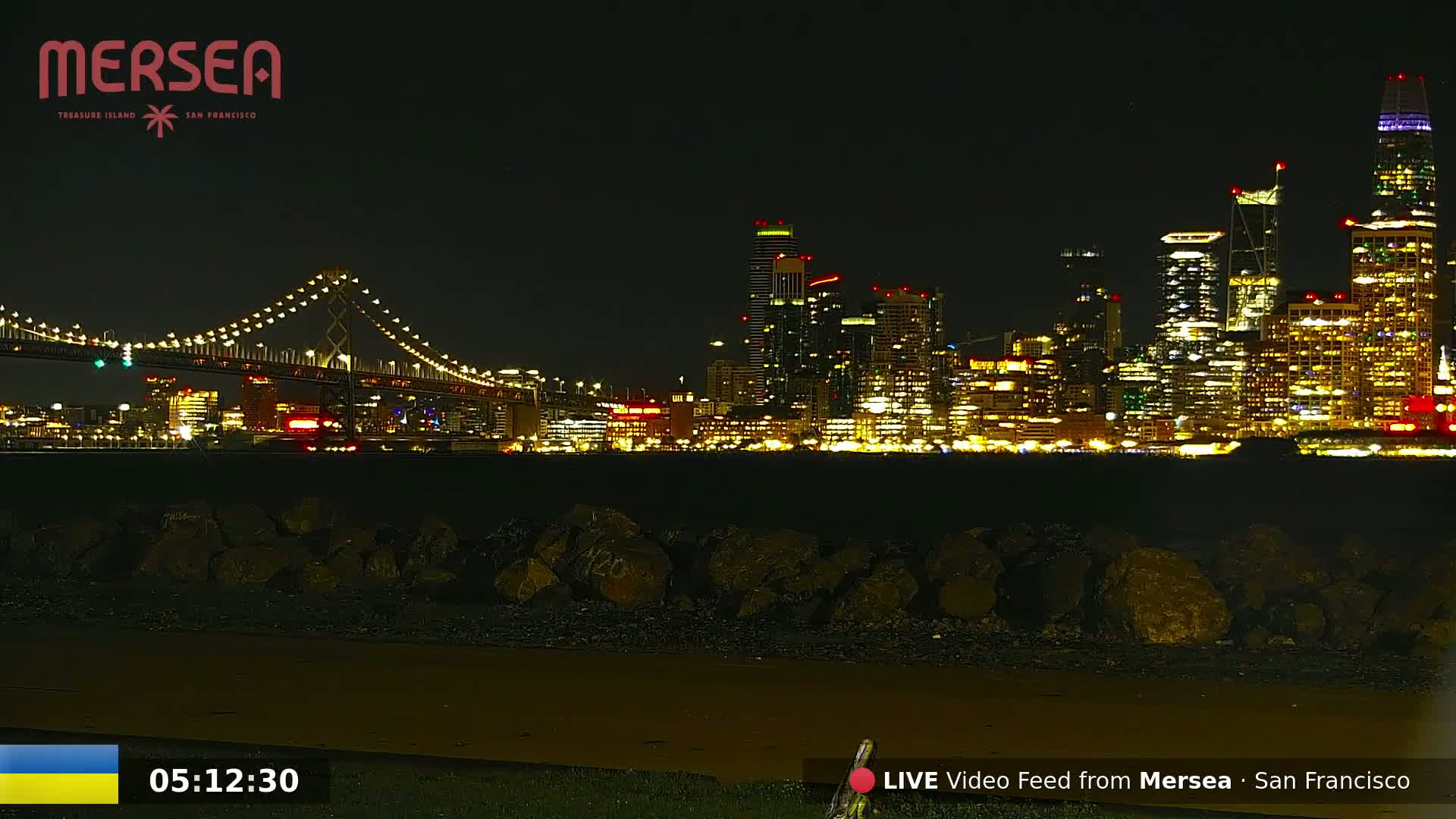A brightly lit cityscape at night is seen across a body of water from a dark shoreline with large rocks, and a lit bridge spans the water in the foreground.