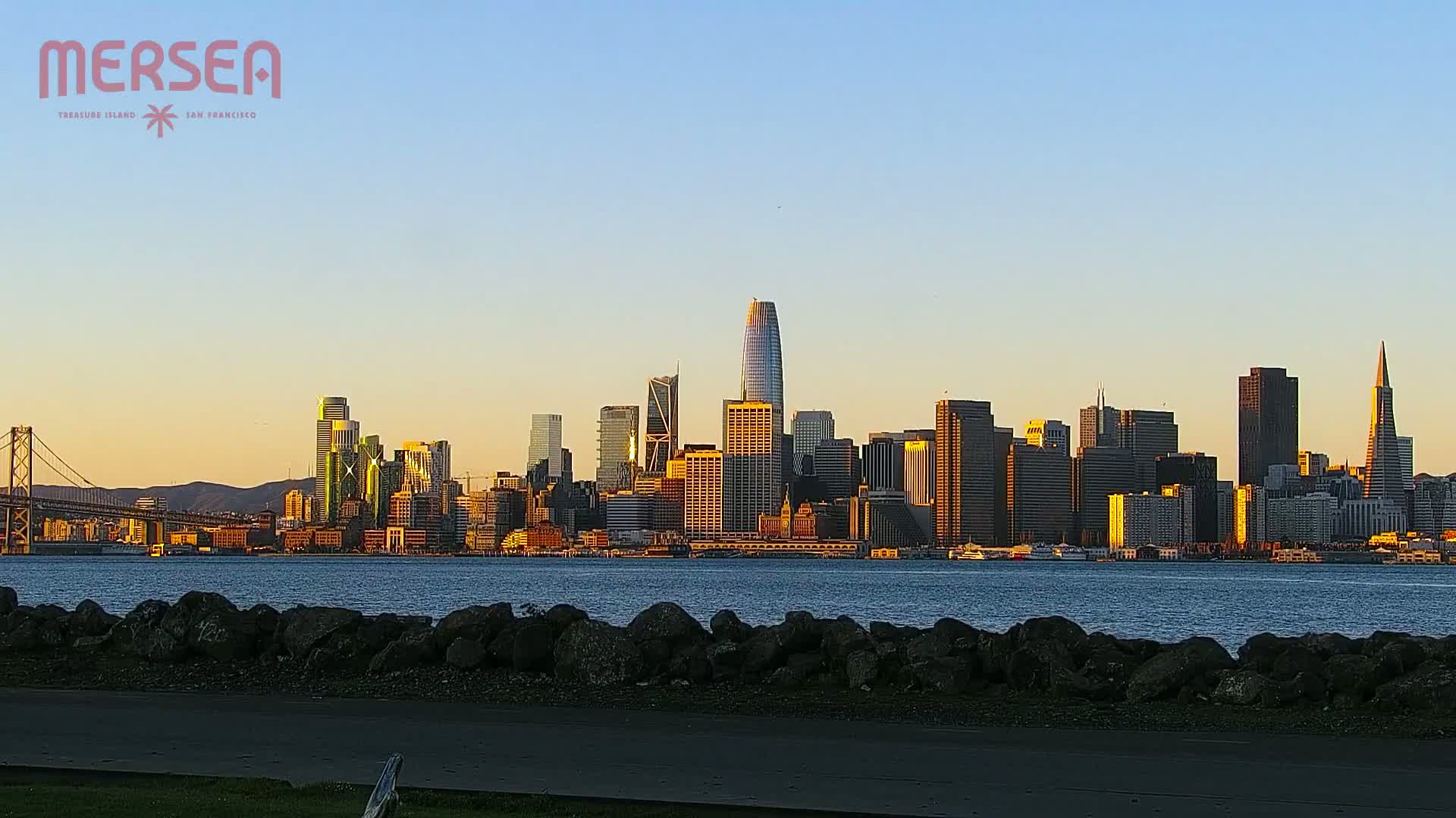 A city skyline, bathed in the golden light of sunset, is seen across a calm body of water from behind a low rock wall.