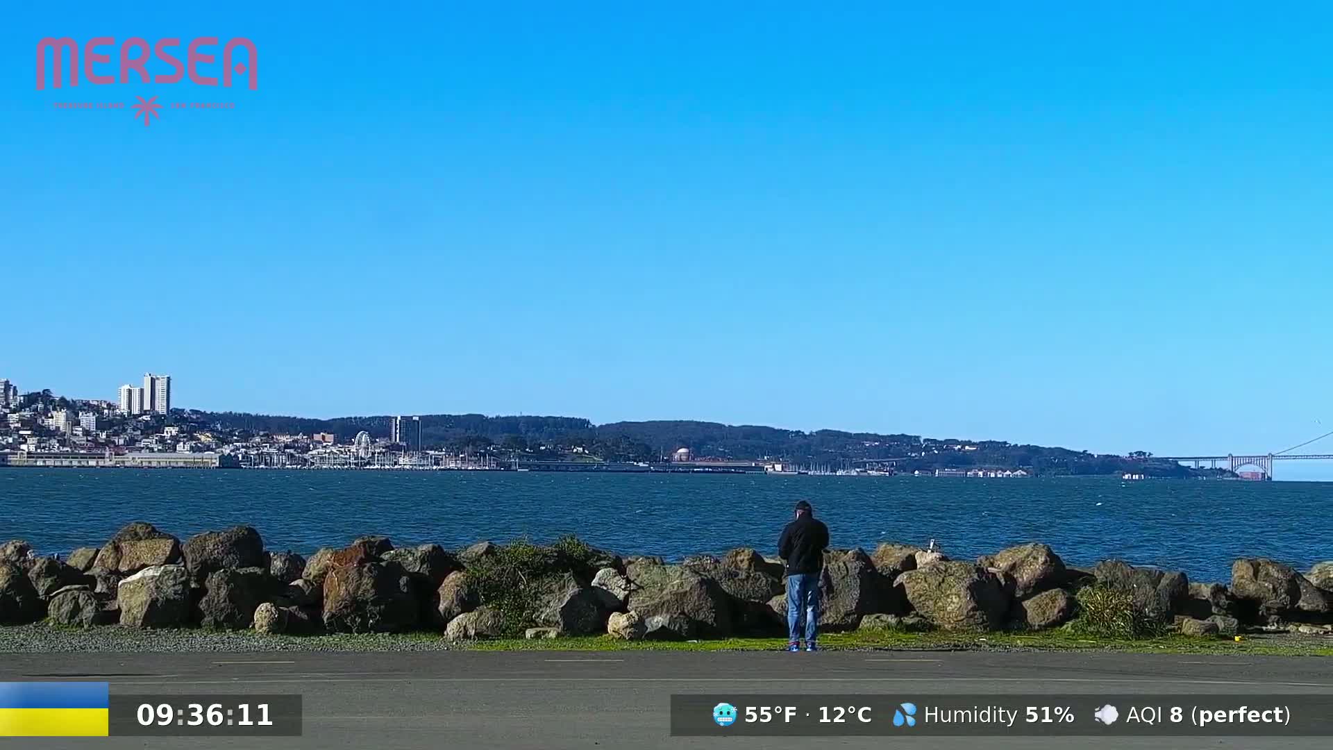 A man stands near a rocky shoreline overlooking a calm body of water, with a city skyline and a bridge visible in the distance under a clear blue sky.