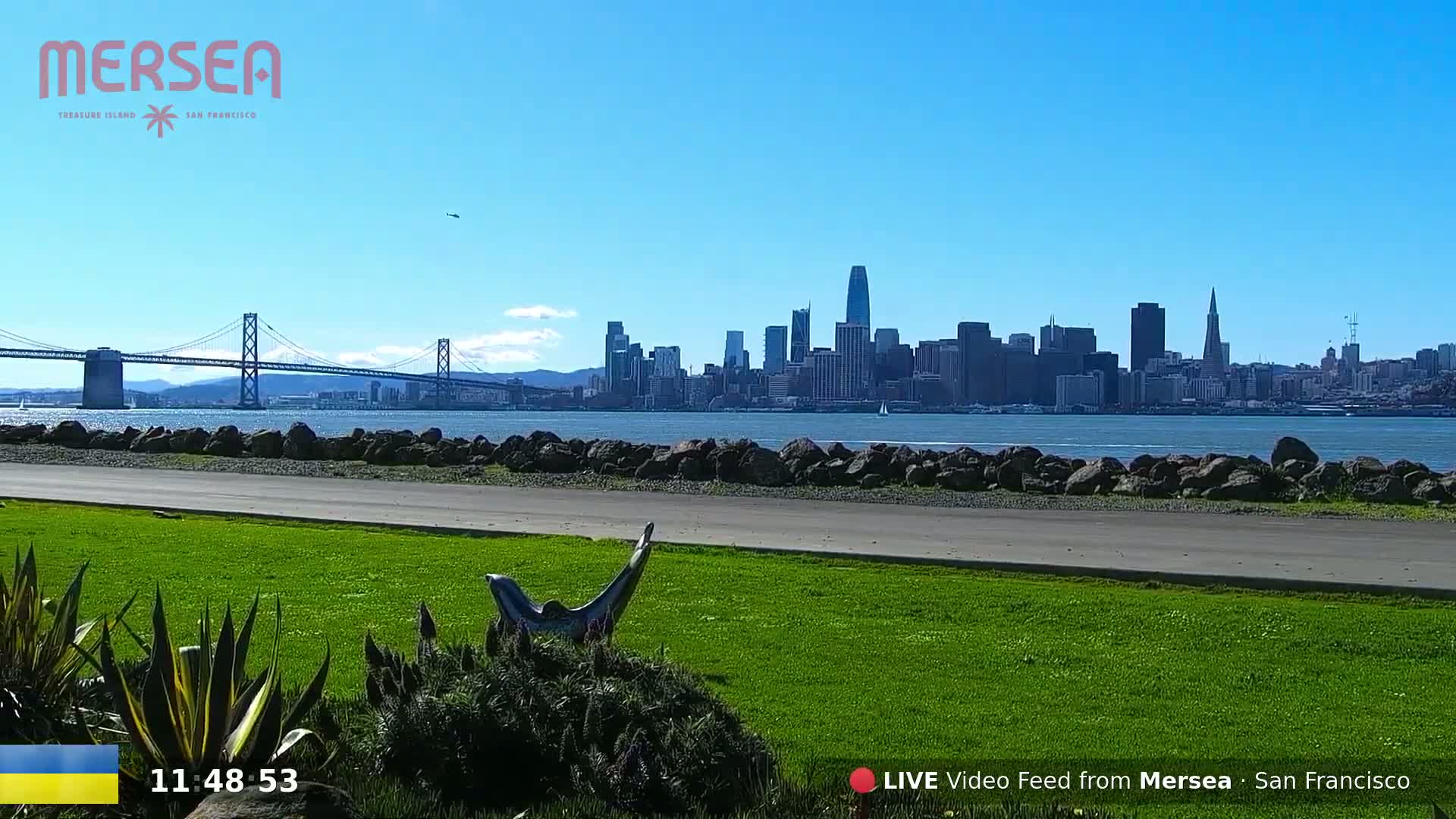 A grassy park area with a sculpture overlooks a body of water, a bridge, and the San Francisco skyline under a clear blue sky.
