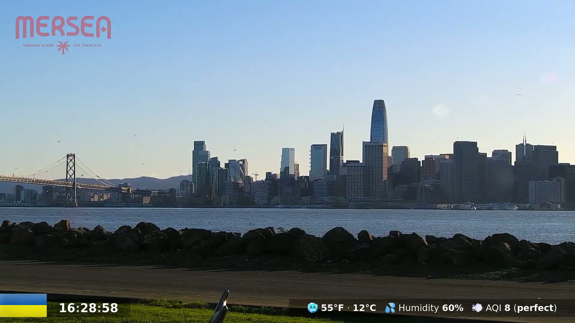 A city skyline, including a suspension bridge, is visible across a calm body of water on a clear, sunny day.