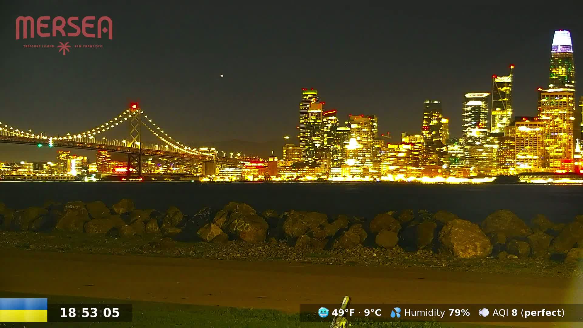 A nighttime view of a city skyline illuminated with lights across a body of water, a bridge in the foreground, and a rocky shoreline in the immediate foreground.
