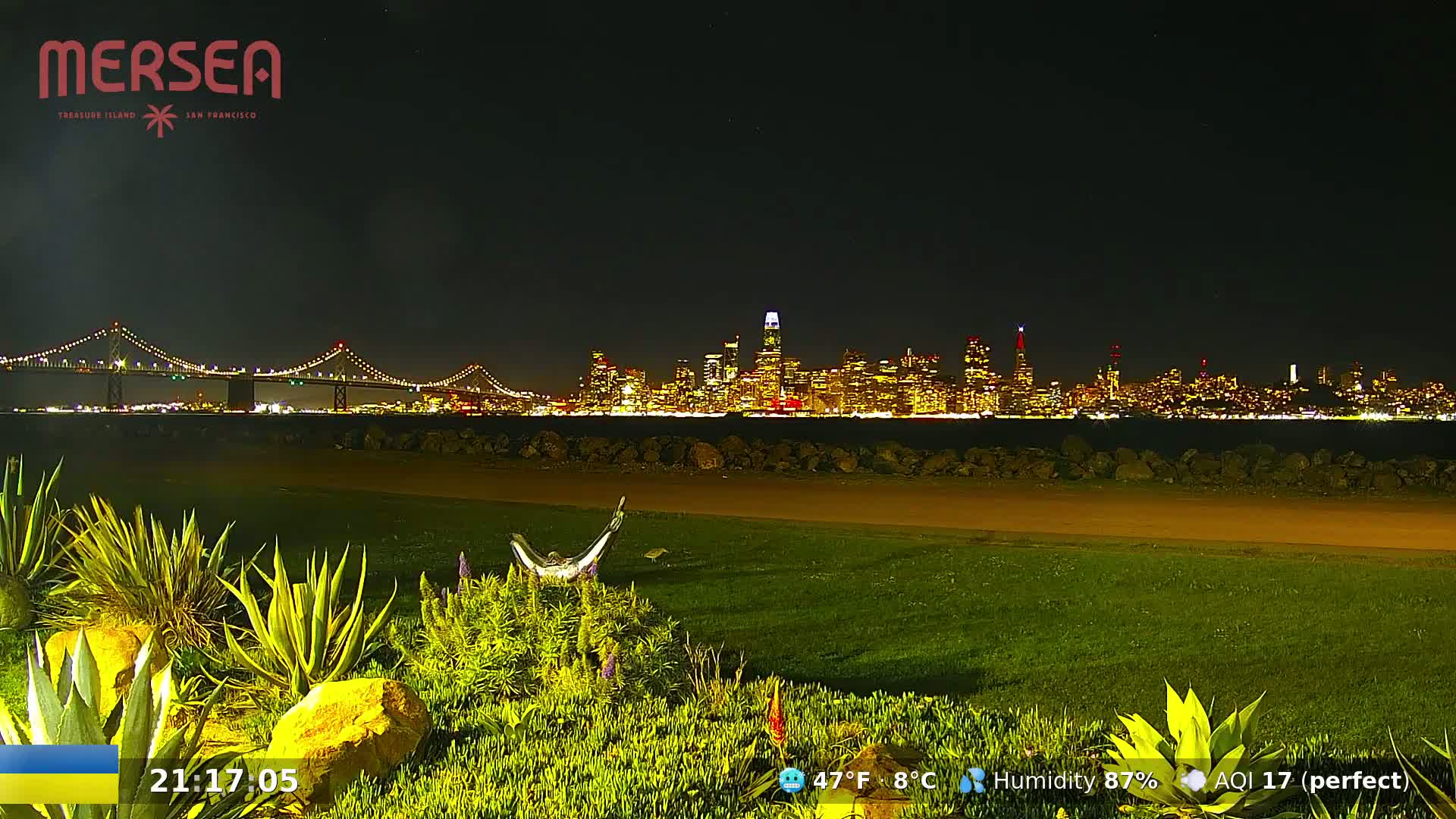 A nighttime view of a city skyline across a body of water, with a bridge in the foreground and landscaped areas in the immediate foreground, under a clear night sky.