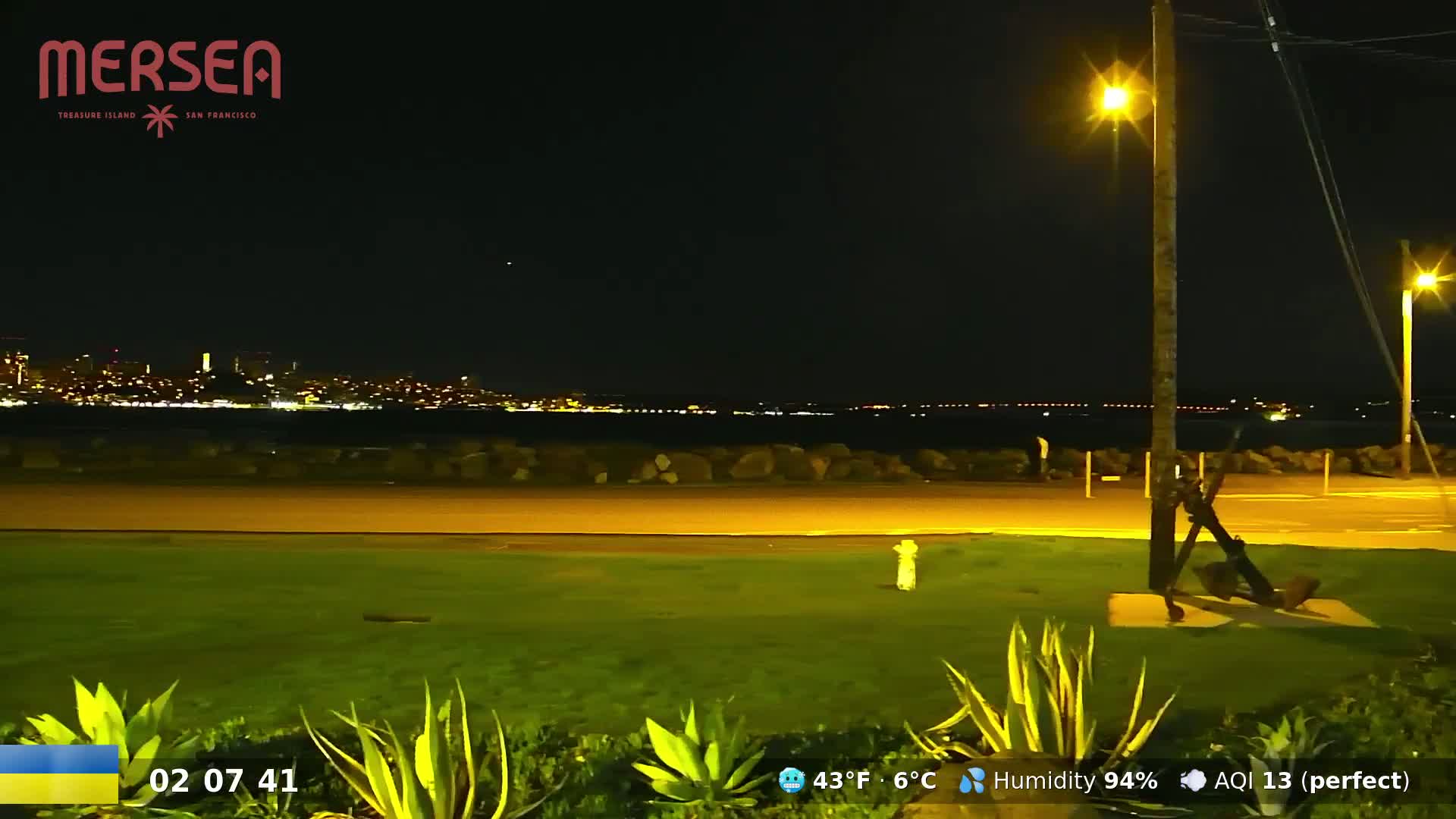 A nighttime view of a city skyline across a body of water, seen from a grassy area with a lamppost and an anchor sculpture in the foreground, under a clear sky.