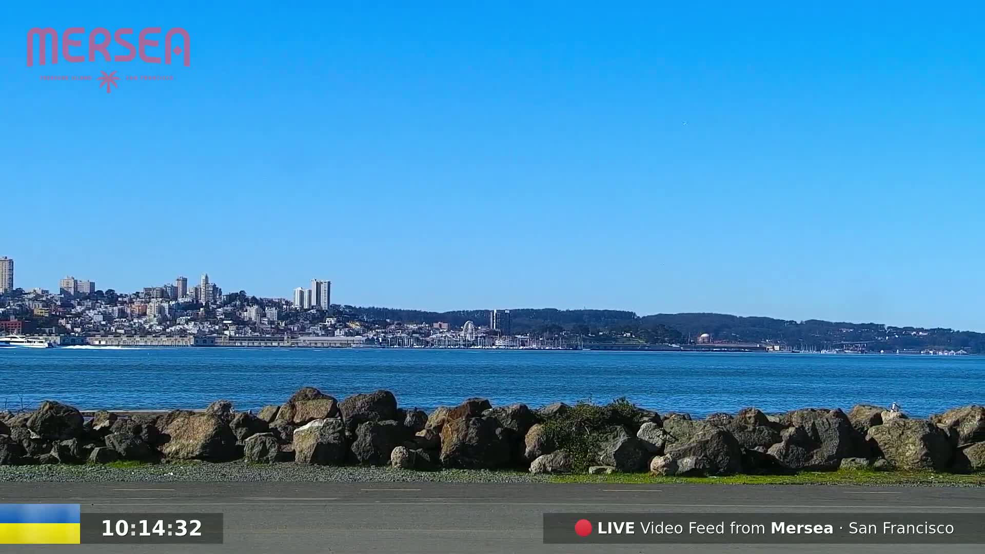 A clear, sunny day shows a waterfront view of a city skyline across a calm body of water, with a rocky shoreline in the foreground.