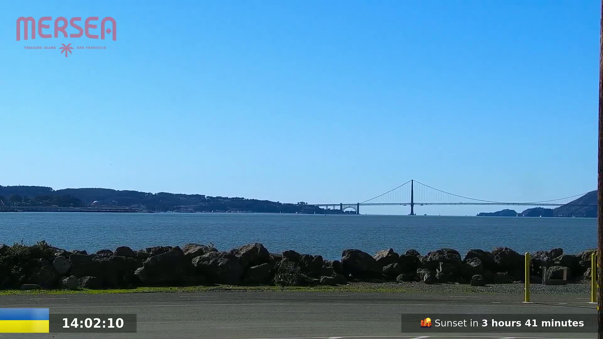 A long suspension bridge is visible in the distance across a calm body of water under a clear blue sky, with dark rocks in the foreground.
