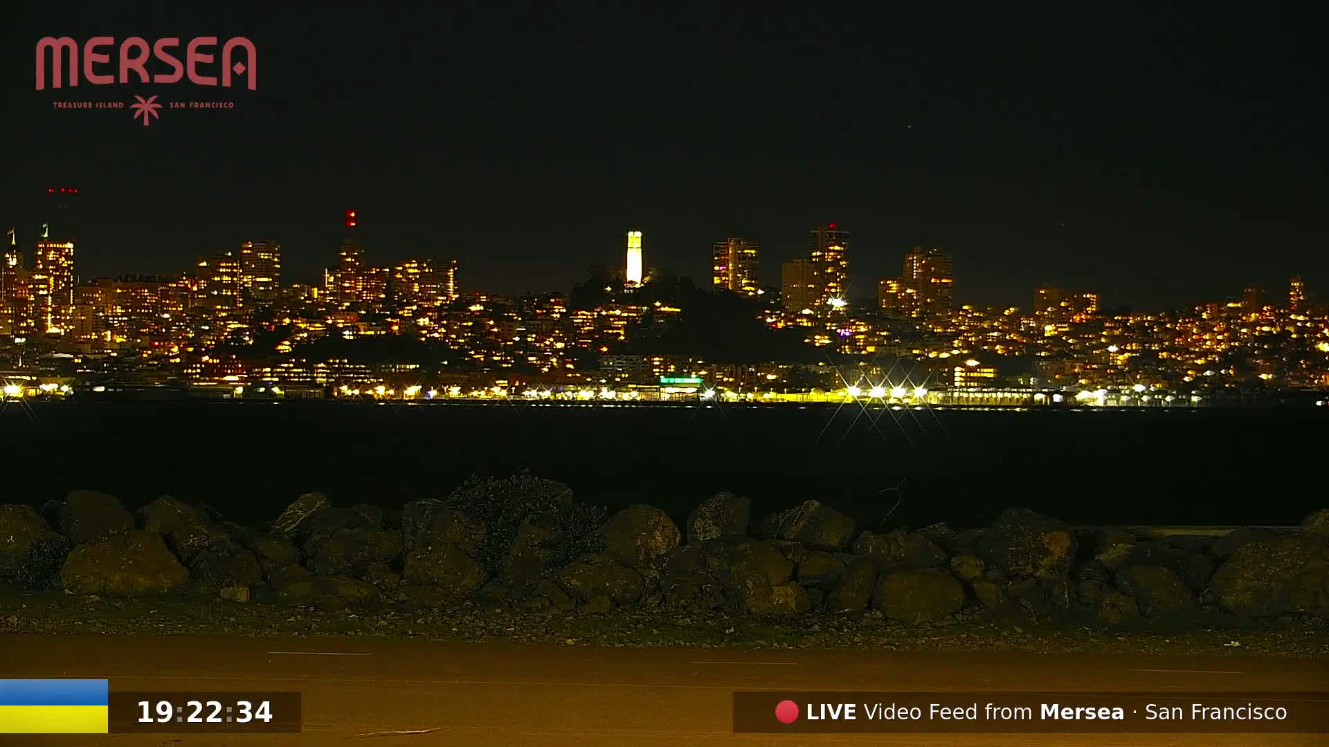 A nighttime view of a city skyline illuminated by numerous lights across a body of water, with a rocky shoreline in the foreground under a clear, dark sky.