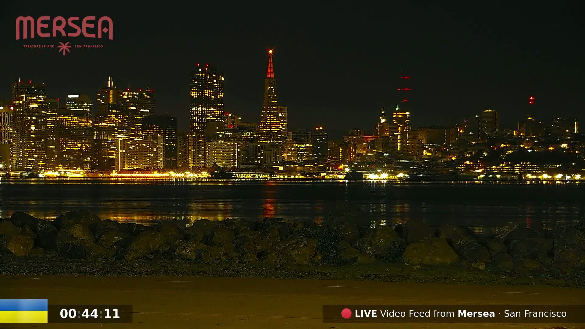 A nighttime view of a city skyline across a body of calm water, with dark rocks in the foreground.