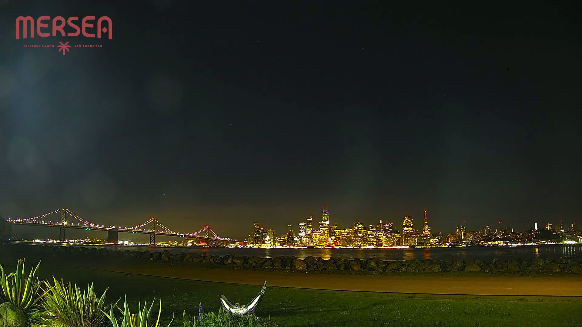 A nighttime view of a city skyline across a body of water, with a bridge in the foreground under a clear, dark sky.