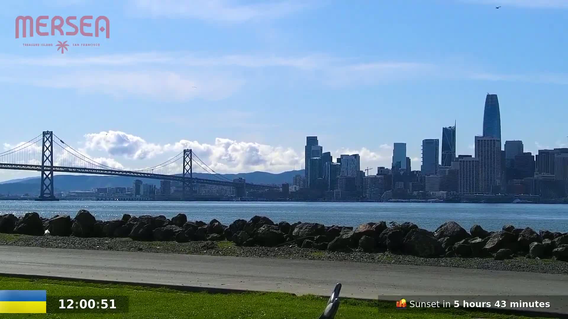 A suspension bridge spans a body of water, with a city skyline and partly cloudy skies in the background.