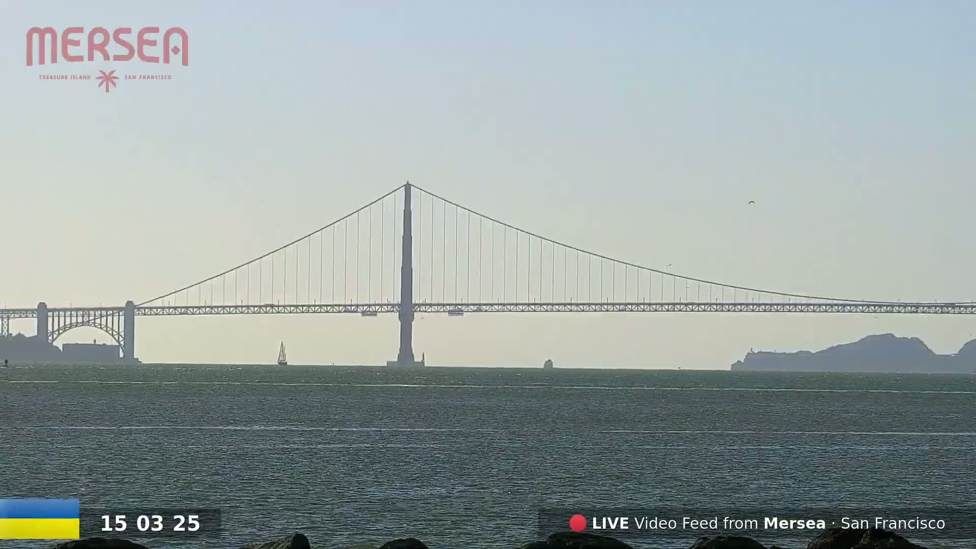 A long suspension bridge spans a calm body of water under a hazy sky.