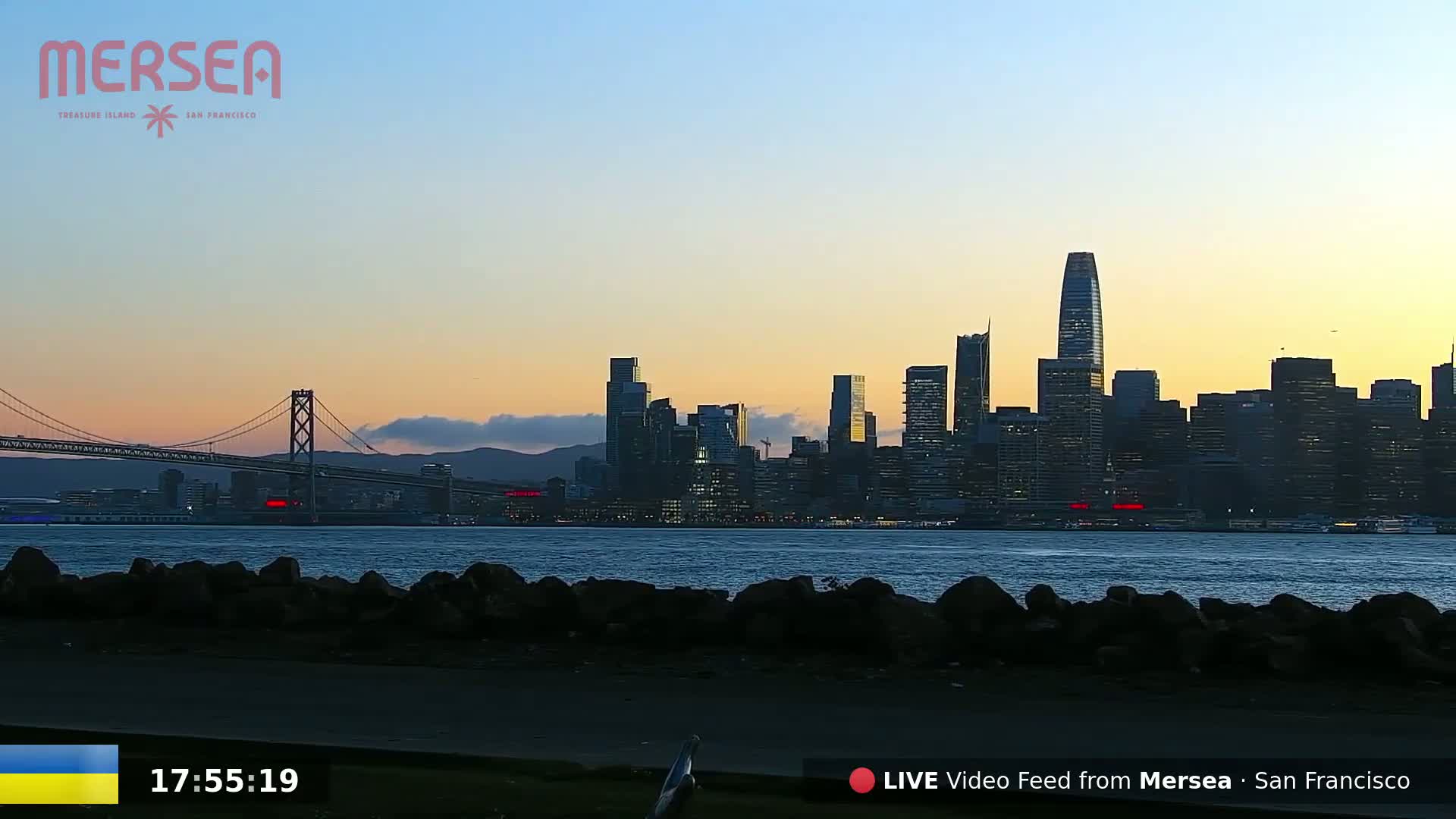 A city skyline at sunset is seen across a body of water from a rocky shoreline.