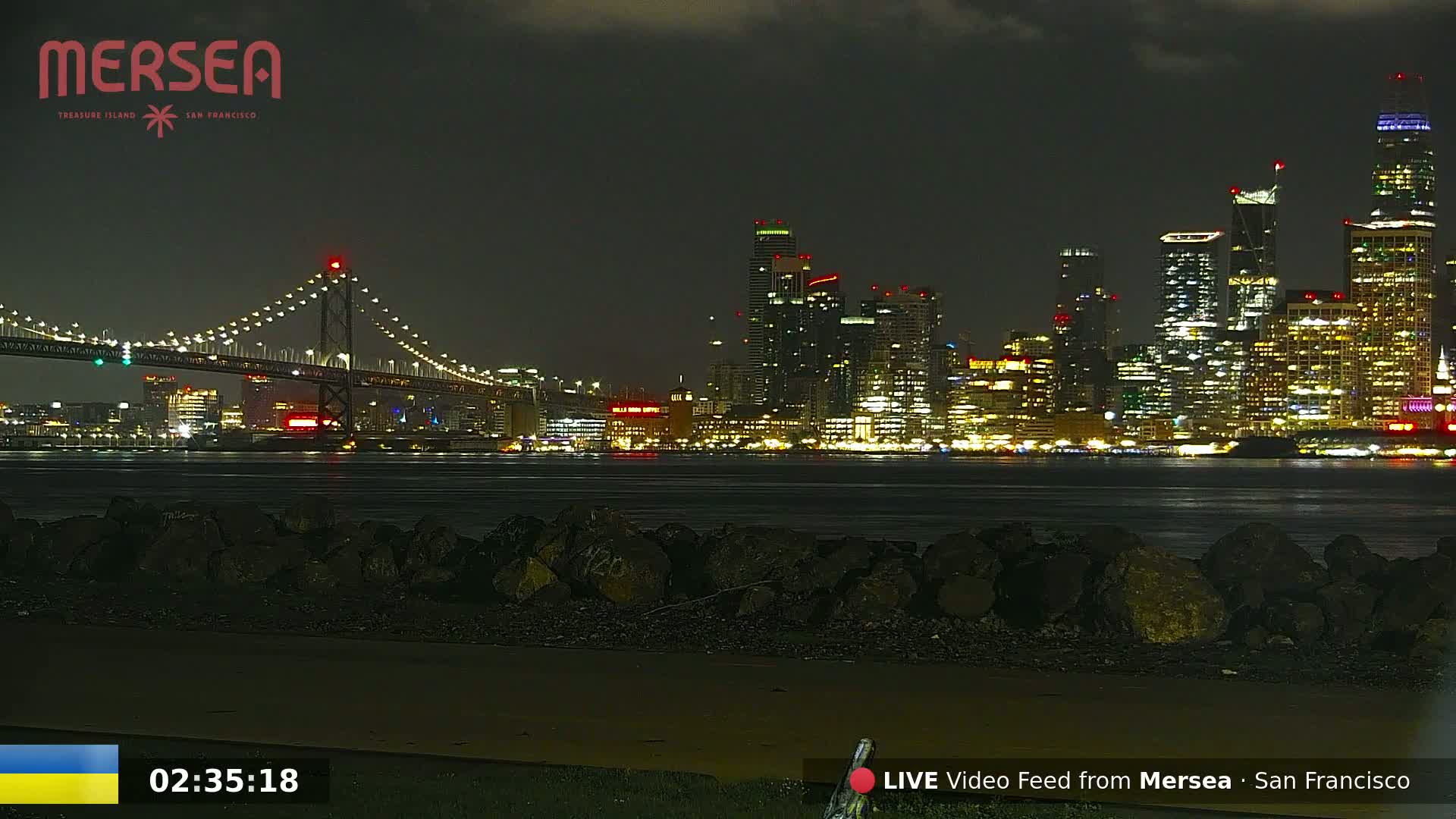 A nighttime view of a city skyline with brightly lit buildings across a body of water, a suspension bridge in the foreground, and a rocky shoreline in the immediate foreground.