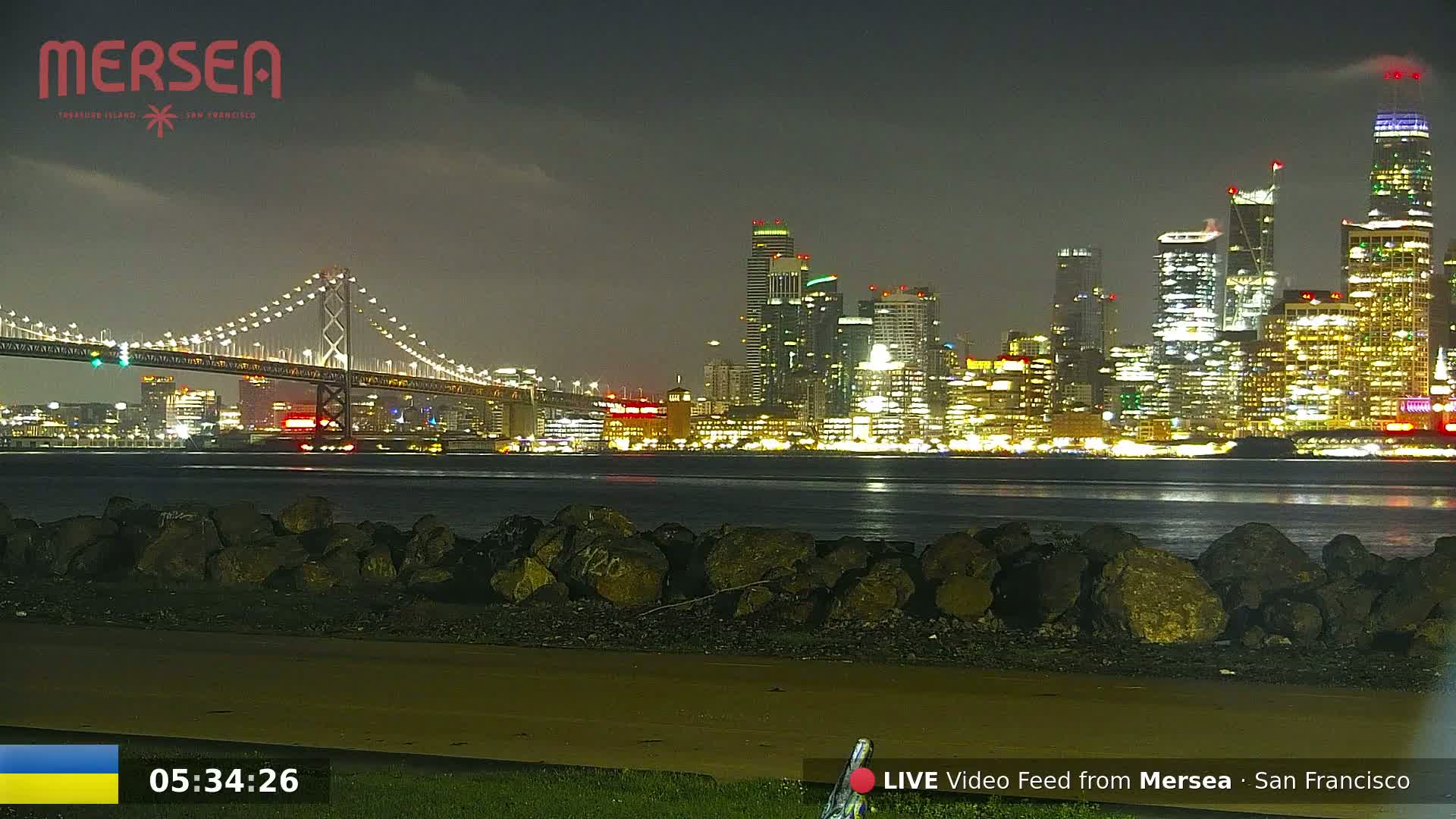 A nighttime view of a lit-up city skyline across a calm body of water, with a bridge stretching into the distance under a mostly clear sky.