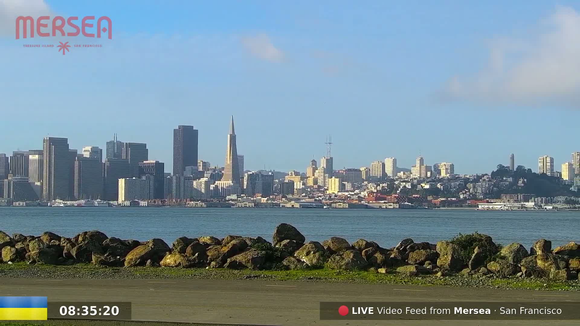 A clear, sunny day reveals a cityscape across a calm body of water, viewed from a rocky shoreline.