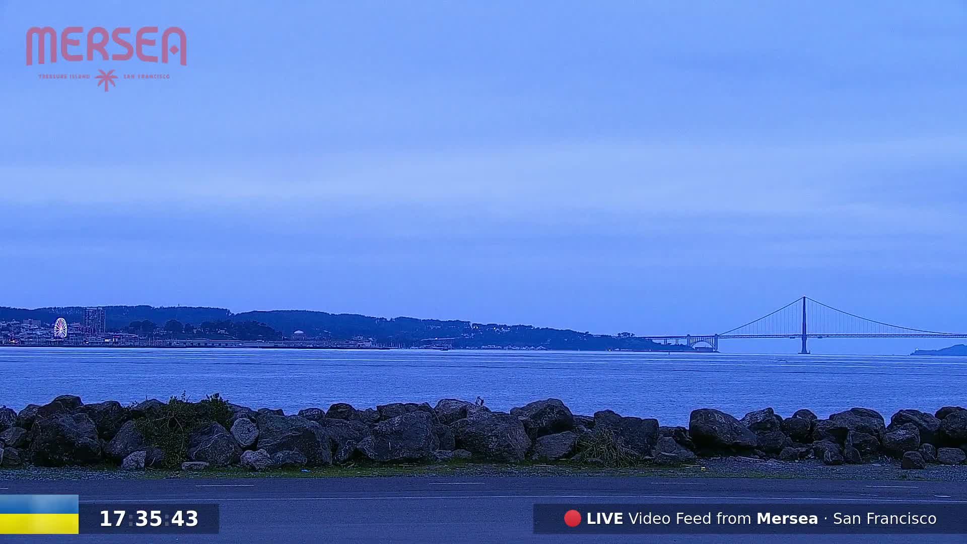 A calm body of water with a distant suspension bridge under a twilight sky is seen behind a low rock wall.