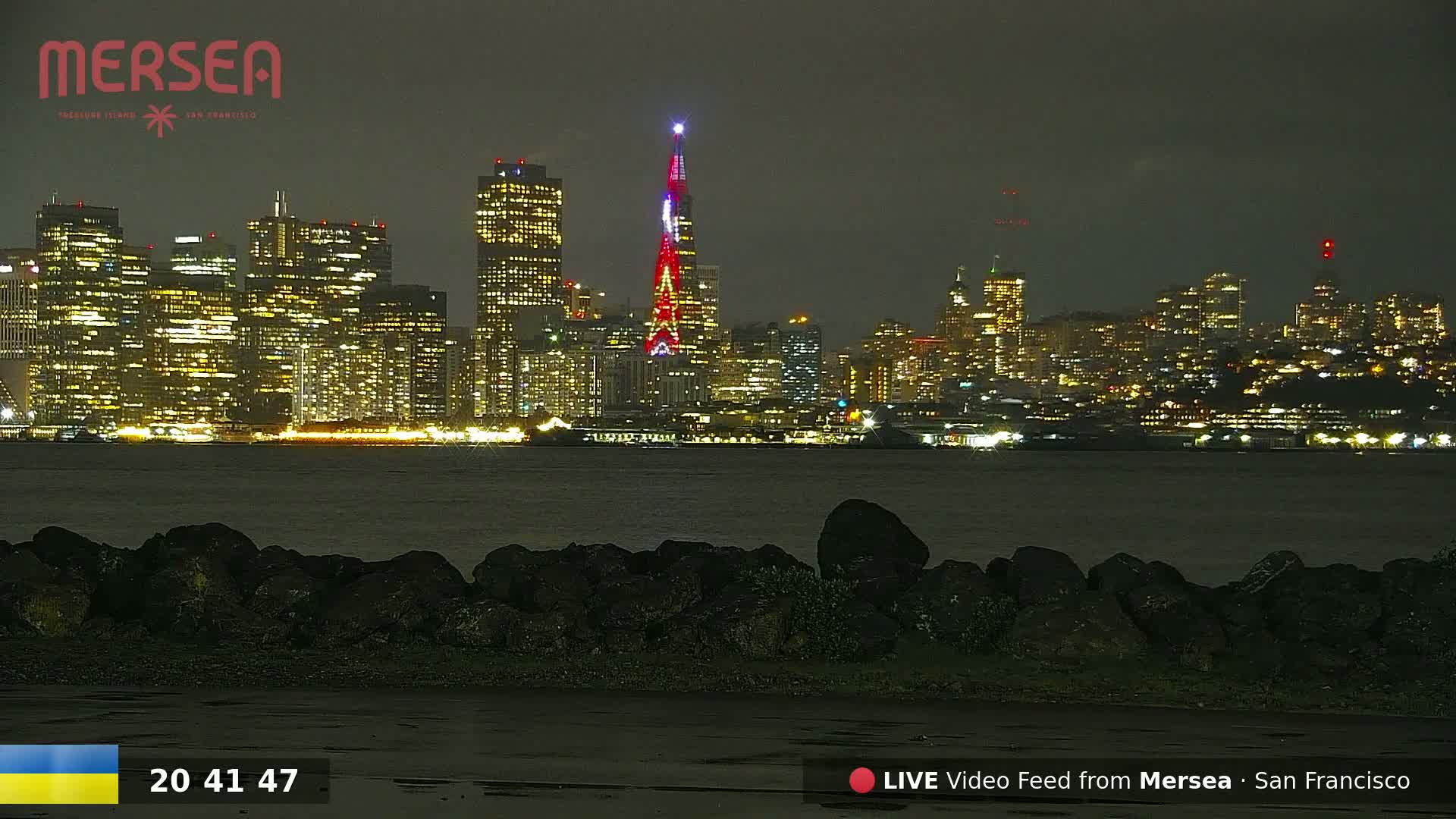 A nighttime view of a city skyline across a body of water, illuminated by city lights, with dark rocks in the foreground.