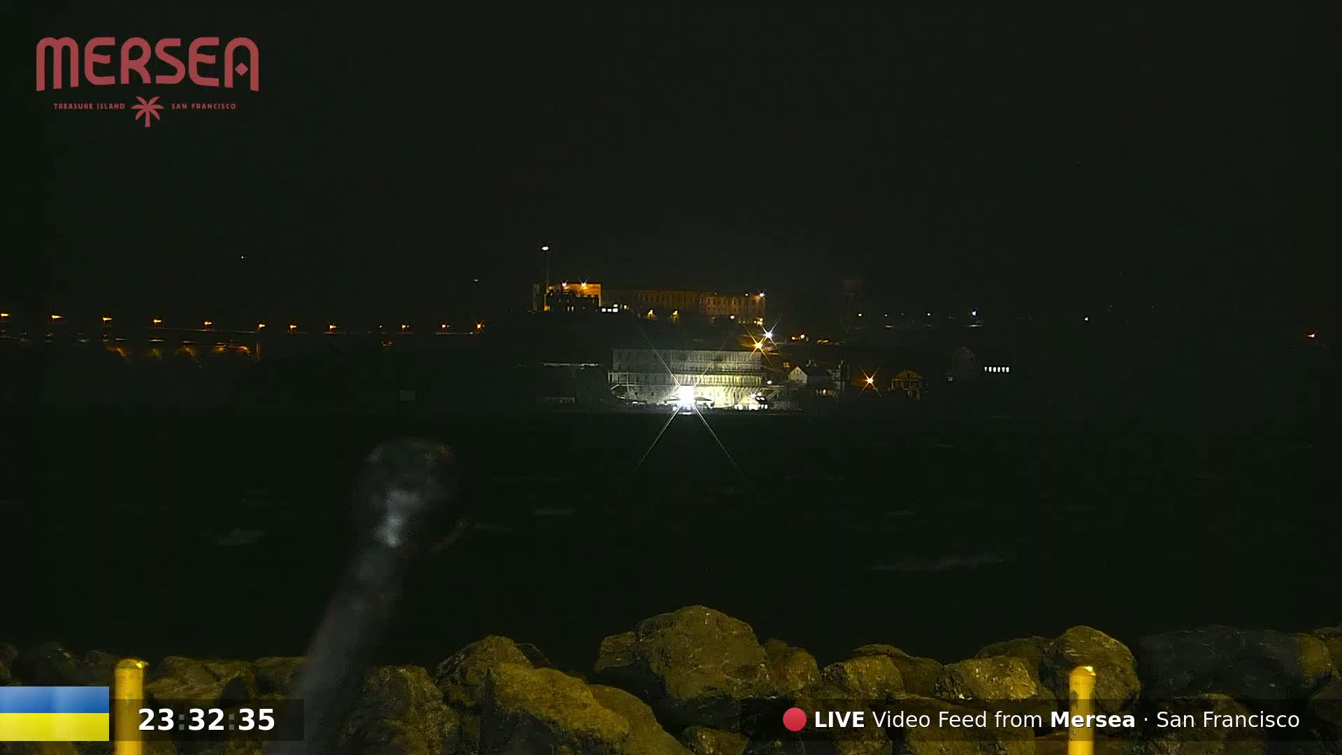 A nighttime view of a building complex illuminated by lights, situated across a dark body of water with rocky shoreline in the foreground.