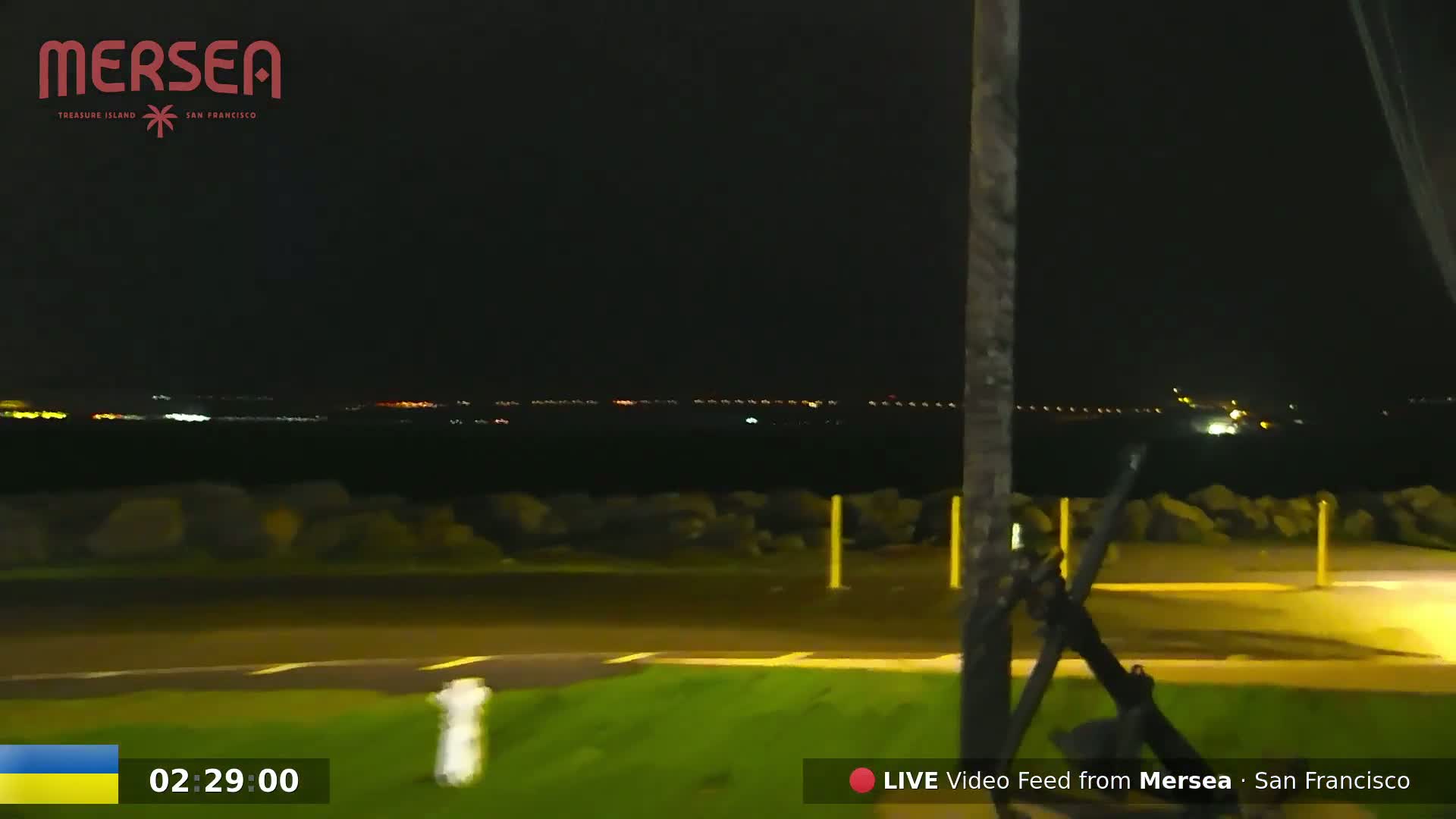 A nighttime view of a dark body of water with distant city lights across it, seen from a grassy area near a roadway.