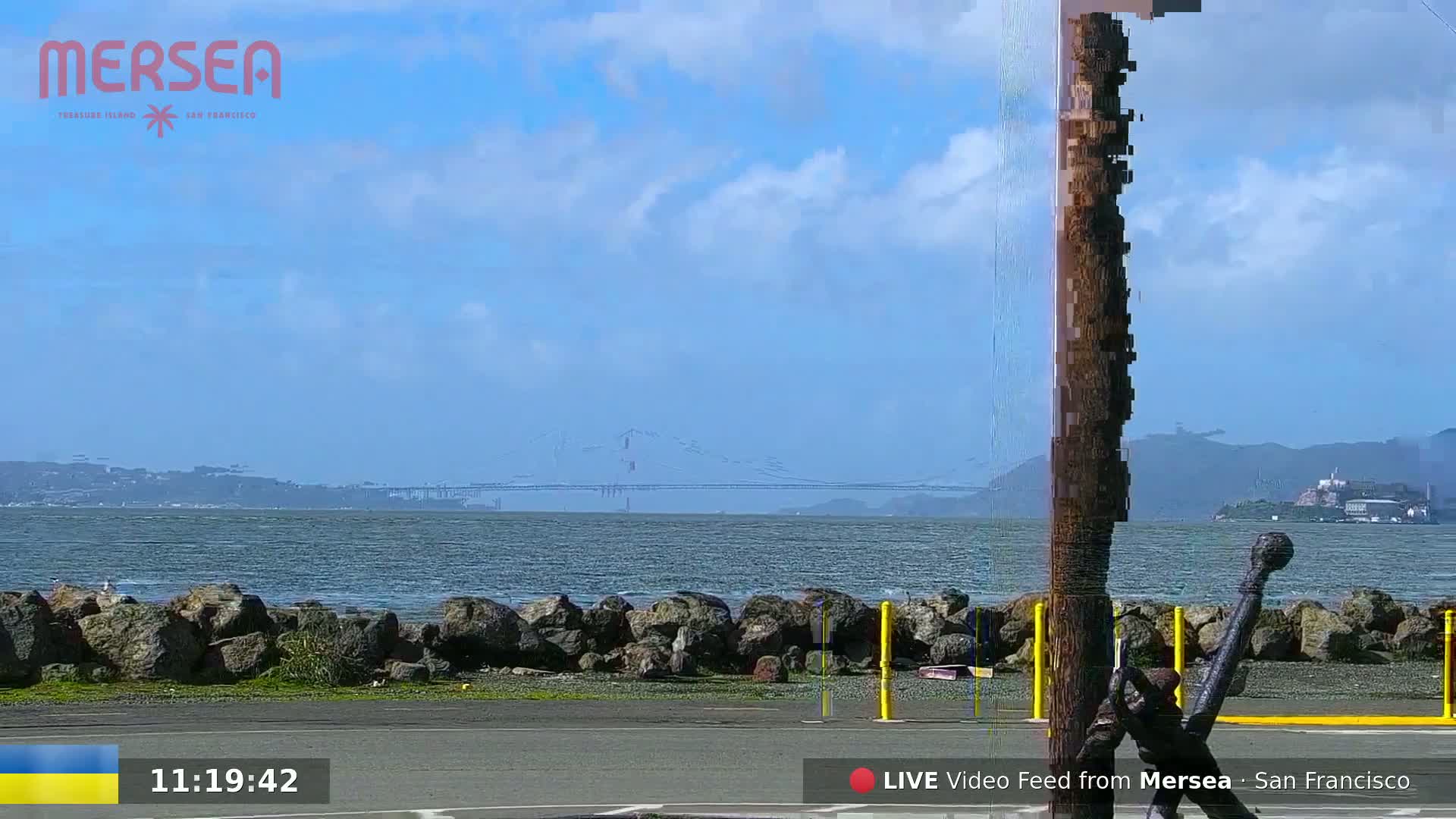 A partly cloudy sky overlooks a body of water with a distant bridge and Alcatraz Island visible under a mostly sunny sky, a rocky shoreline in the foreground, and a large anchor near a wooden post.