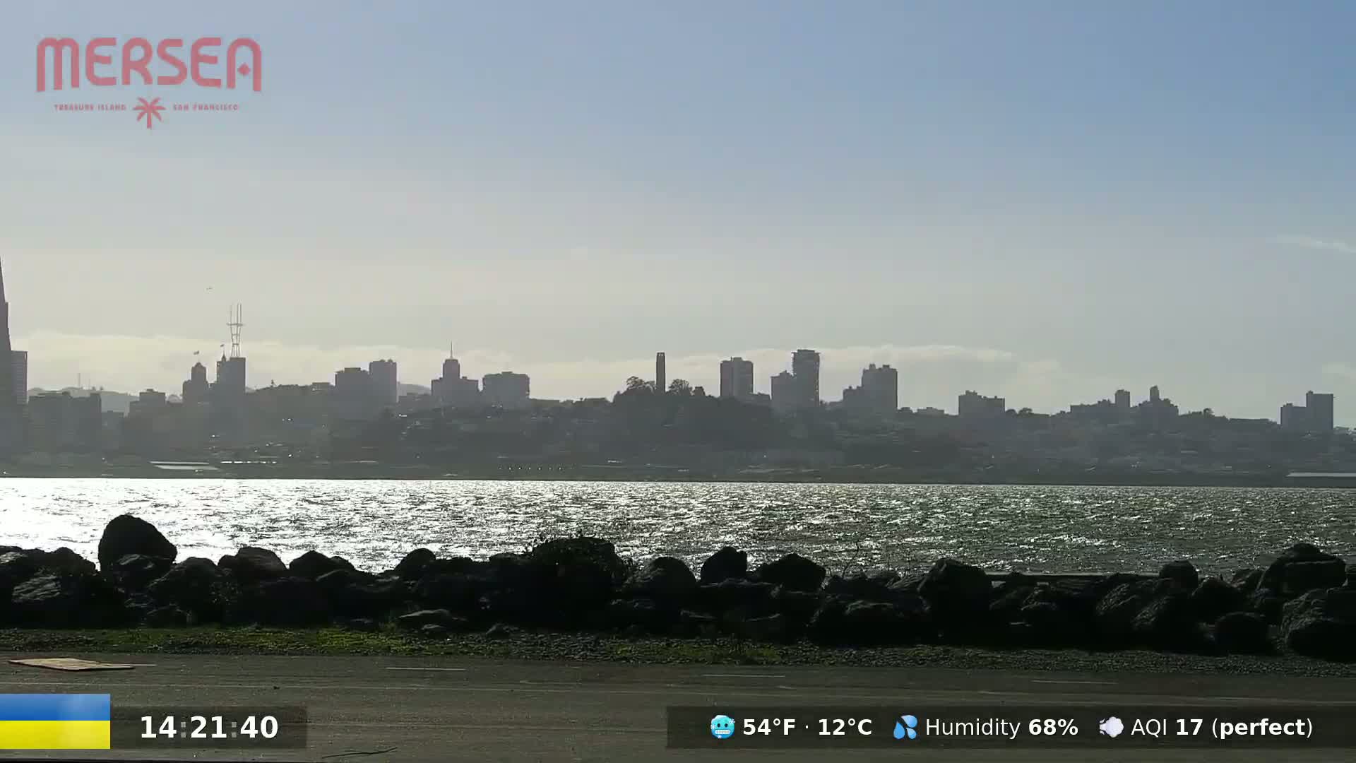 A hazy, sunny day shows a cityscape across a body of water with a rocky shoreline in the foreground.
