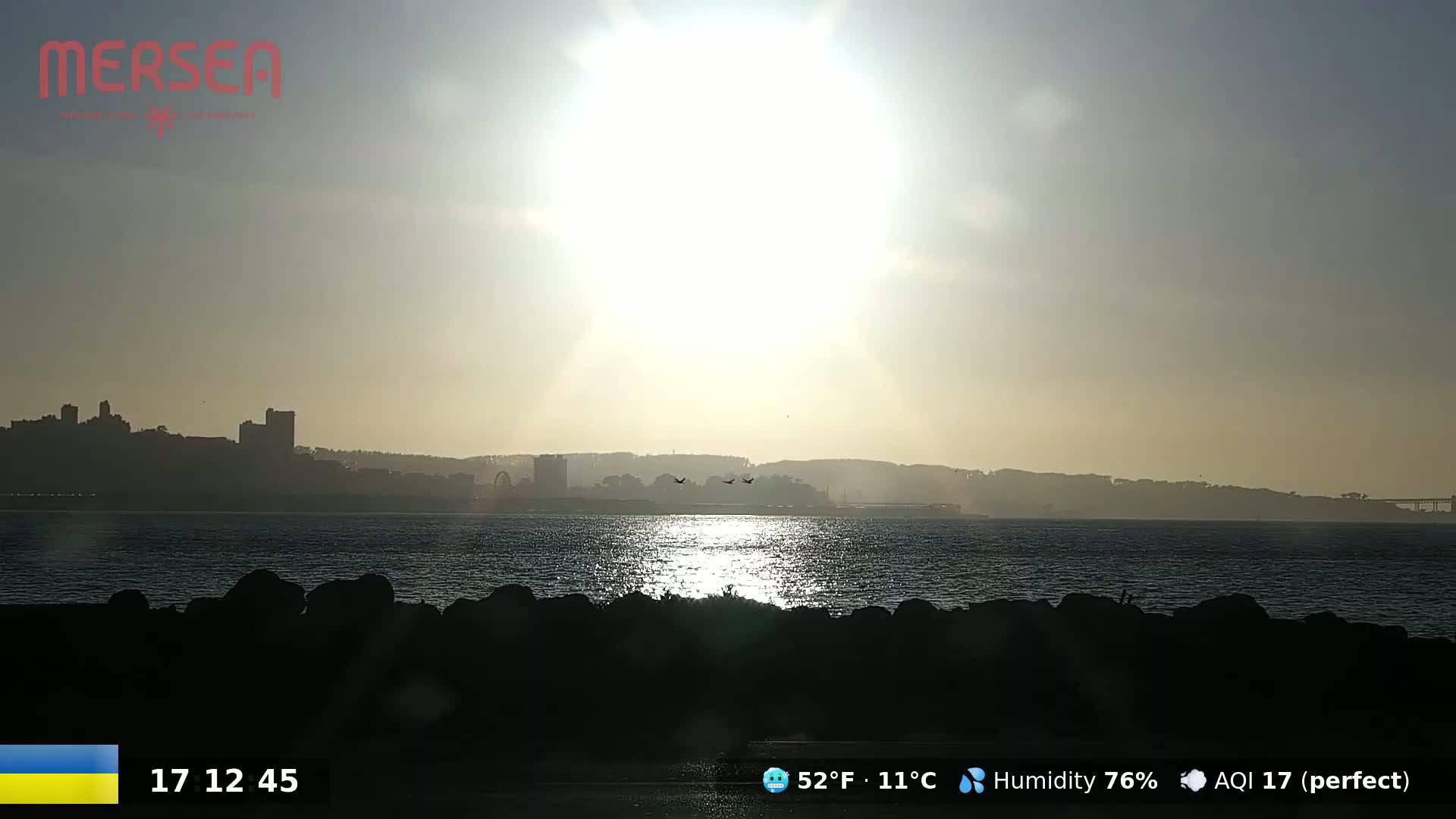 A bright, hazy sun shines over a calm body of water, with a distant city skyline silhouetted against the light.