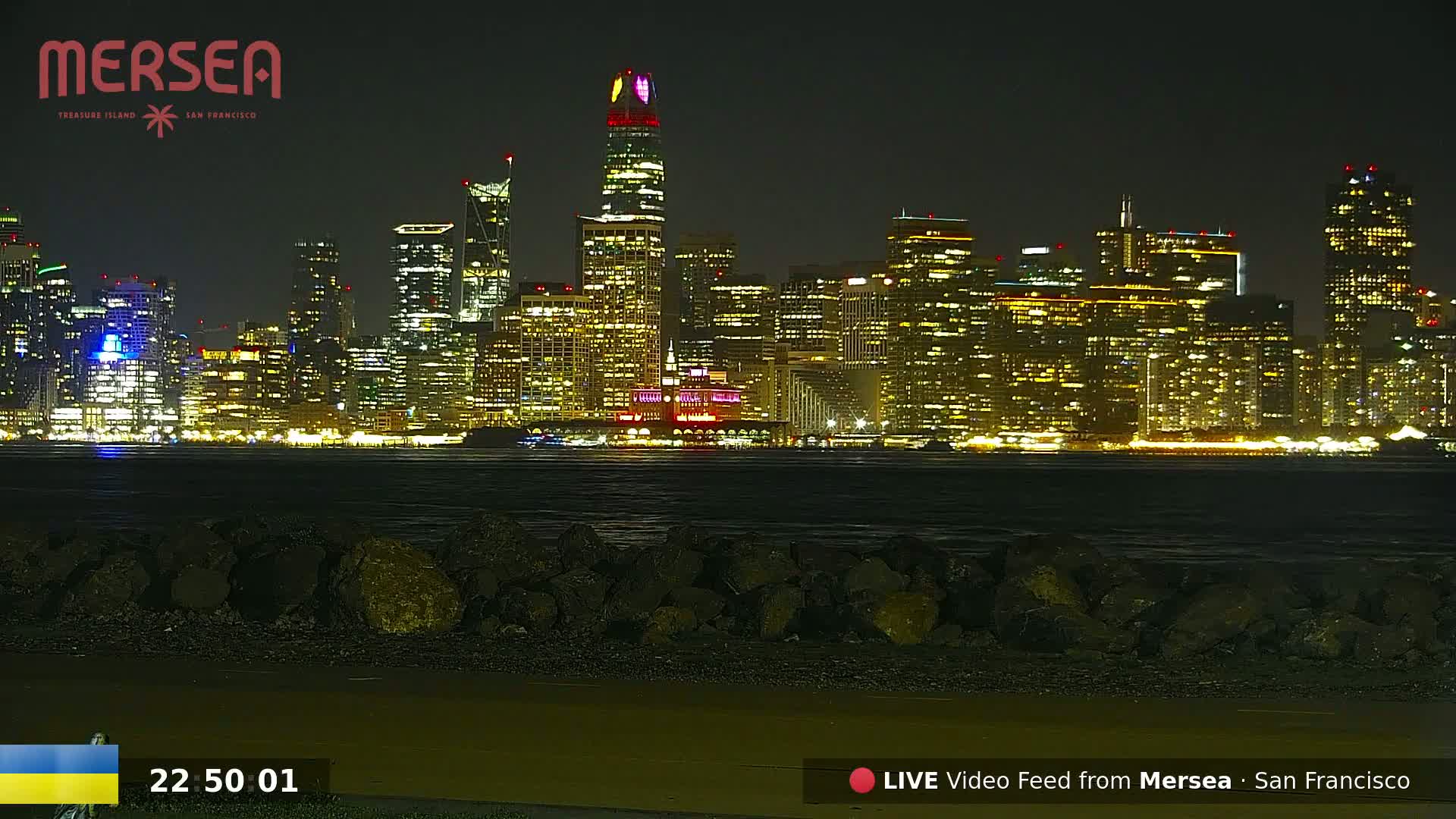 A nighttime view of a city skyline across a dark body of water, with a rocky shoreline in the foreground.