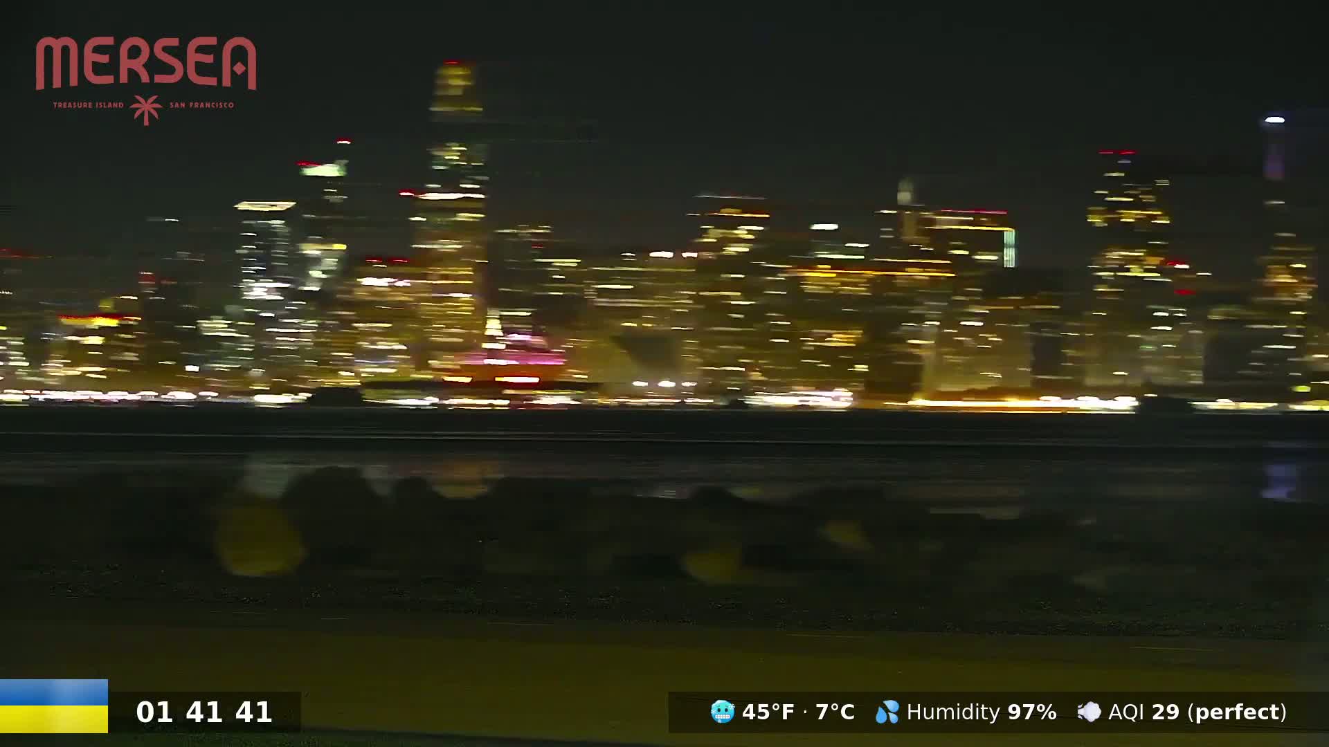 A nighttime, blurry view of a brightly lit city skyline across a dark body of water, under conditions of 45°F (7°C) temperature and 97% humidity.