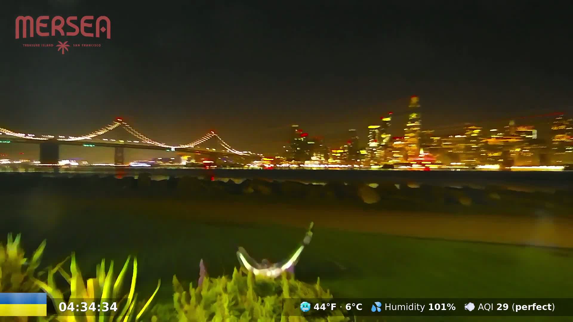 A nighttime view of a city skyline across a body of water, illuminated by city lights, with a bridge in the foreground.