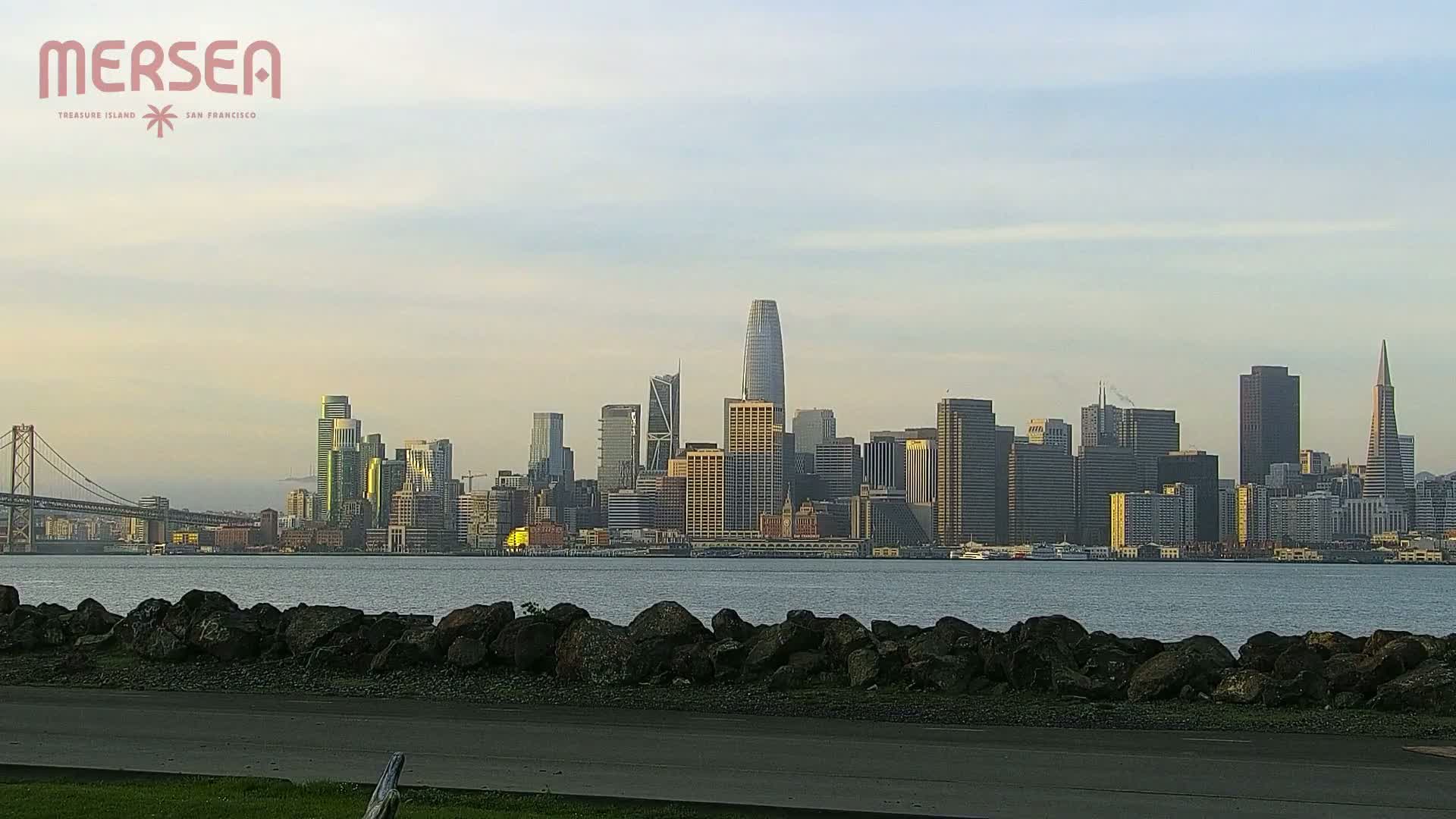 A city skyline with numerous skyscrapers is seen across a body of water under a partly cloudy sky, with a bridge visible to the left.