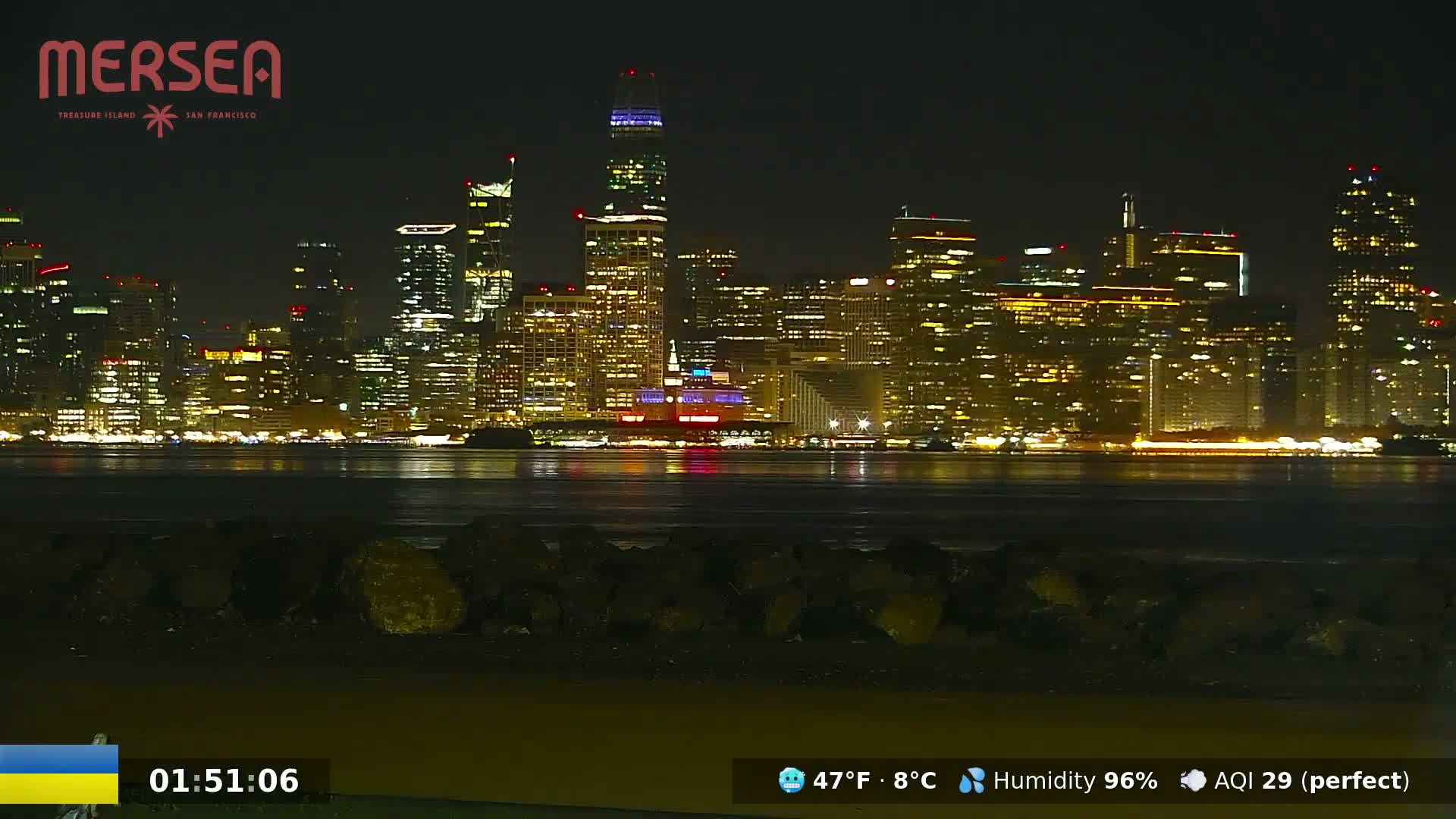 A brightly lit city skyline is seen across a dark body of water at night, under clear skies.