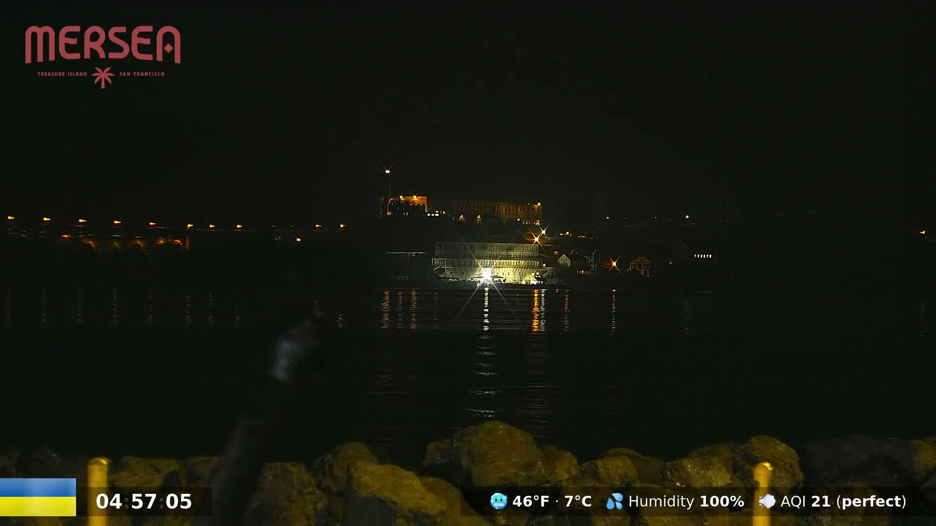A nighttime view across dark water shows a brightly lit building and other structures on a distant shore.