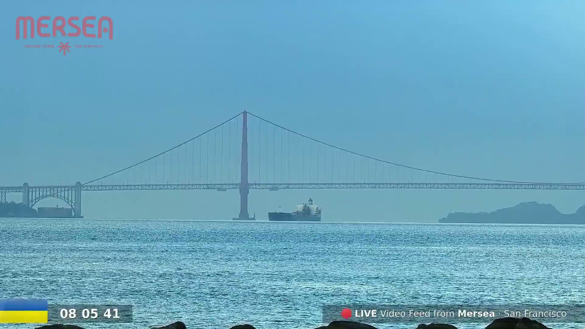 A large cargo ship is sailing beneath the Golden Gate Bridge on a hazy, sunny day.