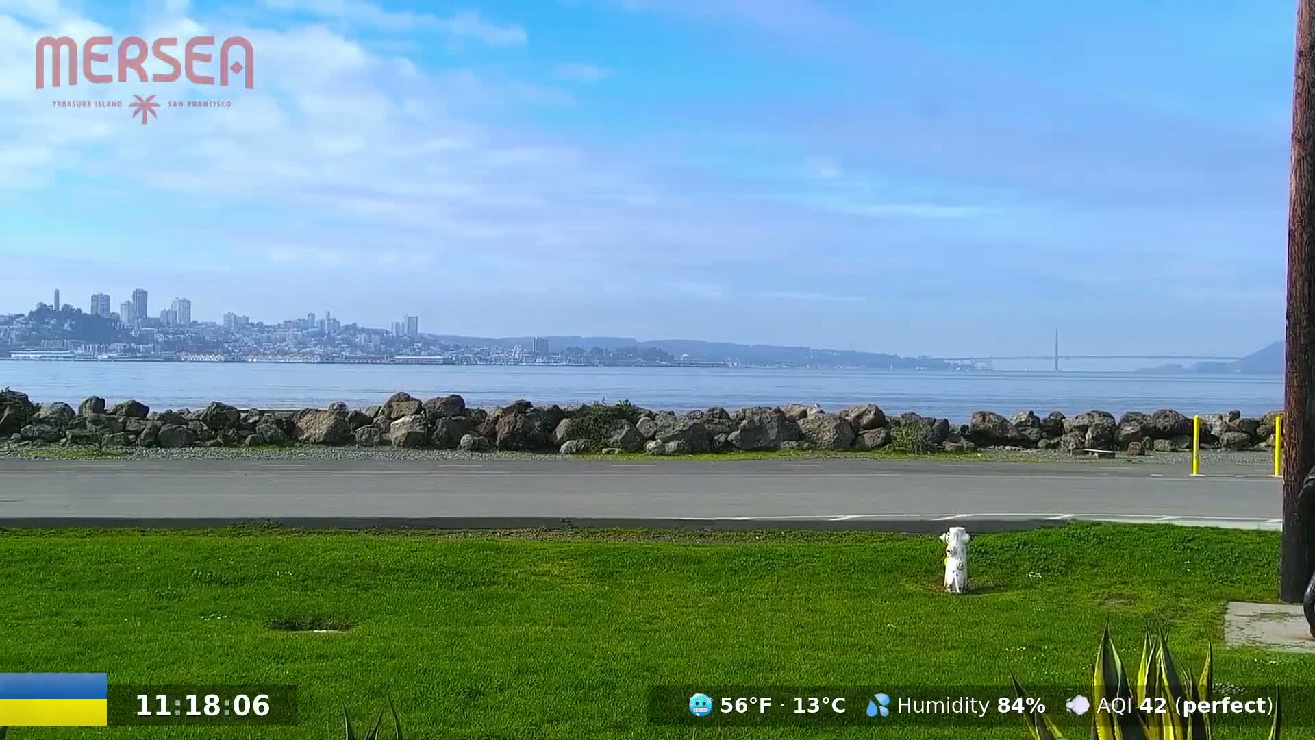 A grassy area with a paved path overlooks a calm body of water under a mostly clear sky, with a distant cityscape visible on the horizon.