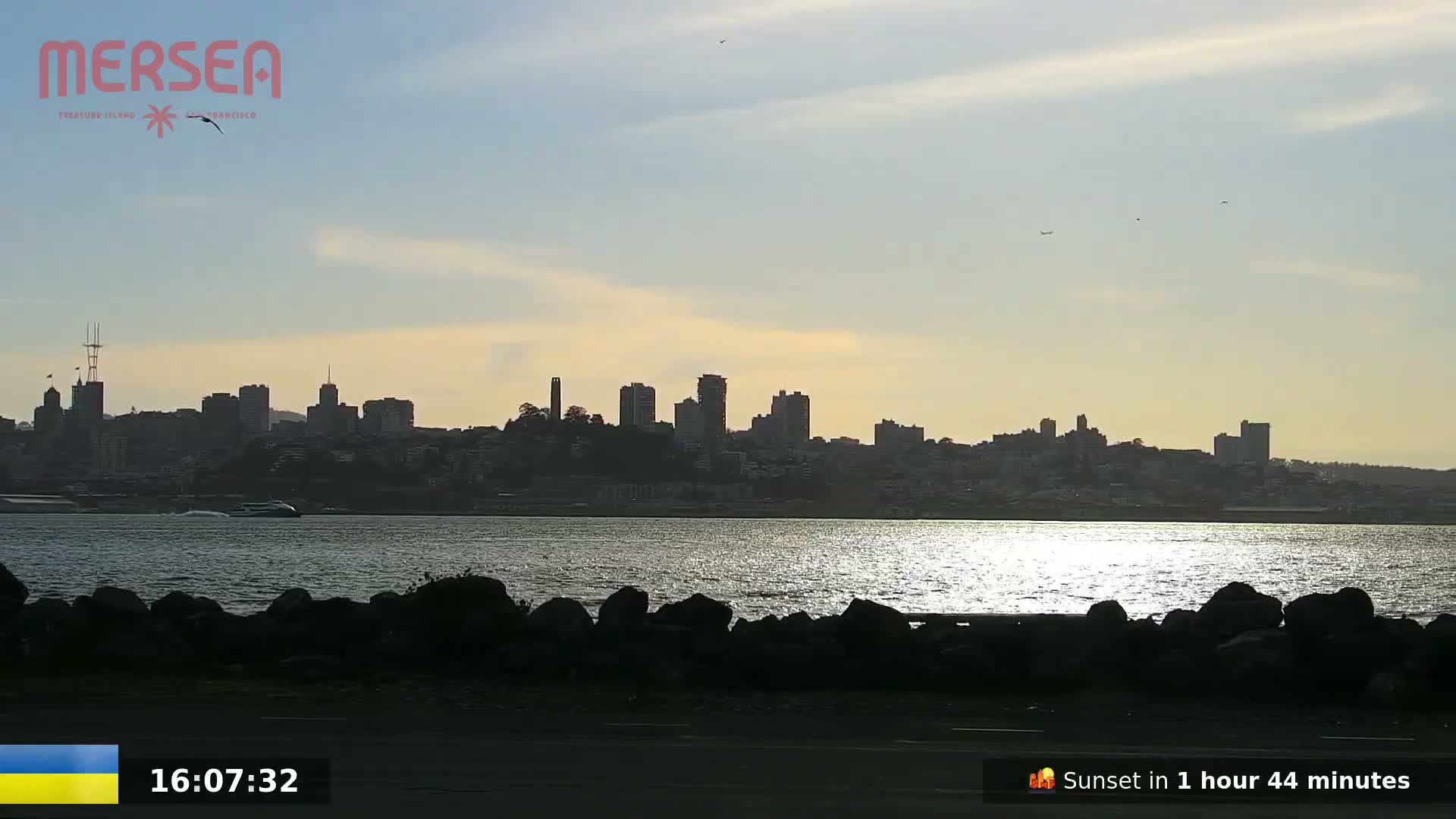 A city skyline is visible across a calm body of water under a partly cloudy sky.