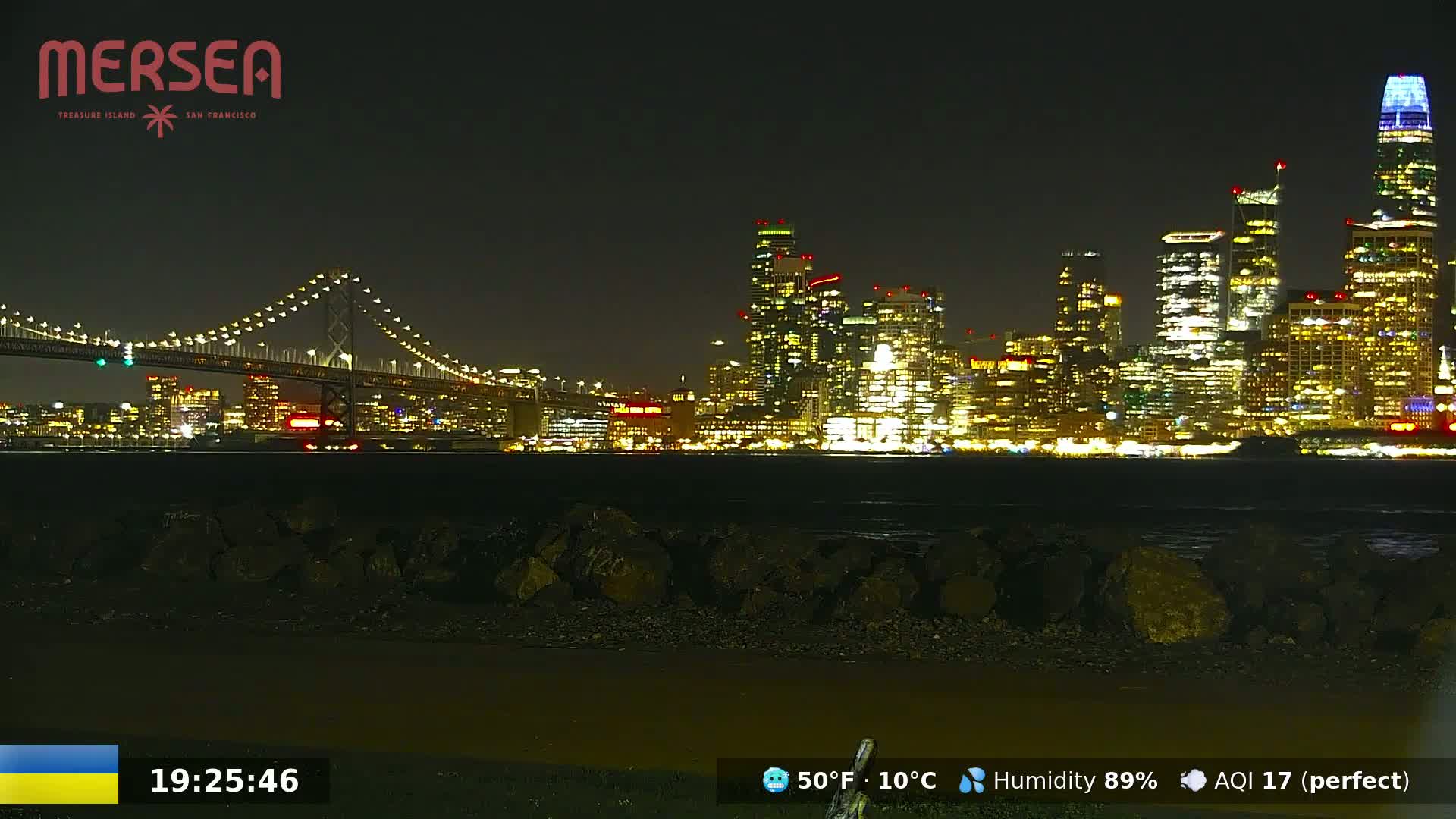 A nighttime view of a city skyline illuminated with bright lights across a body of water, with a bridge in the foreground under a clear, dark sky.