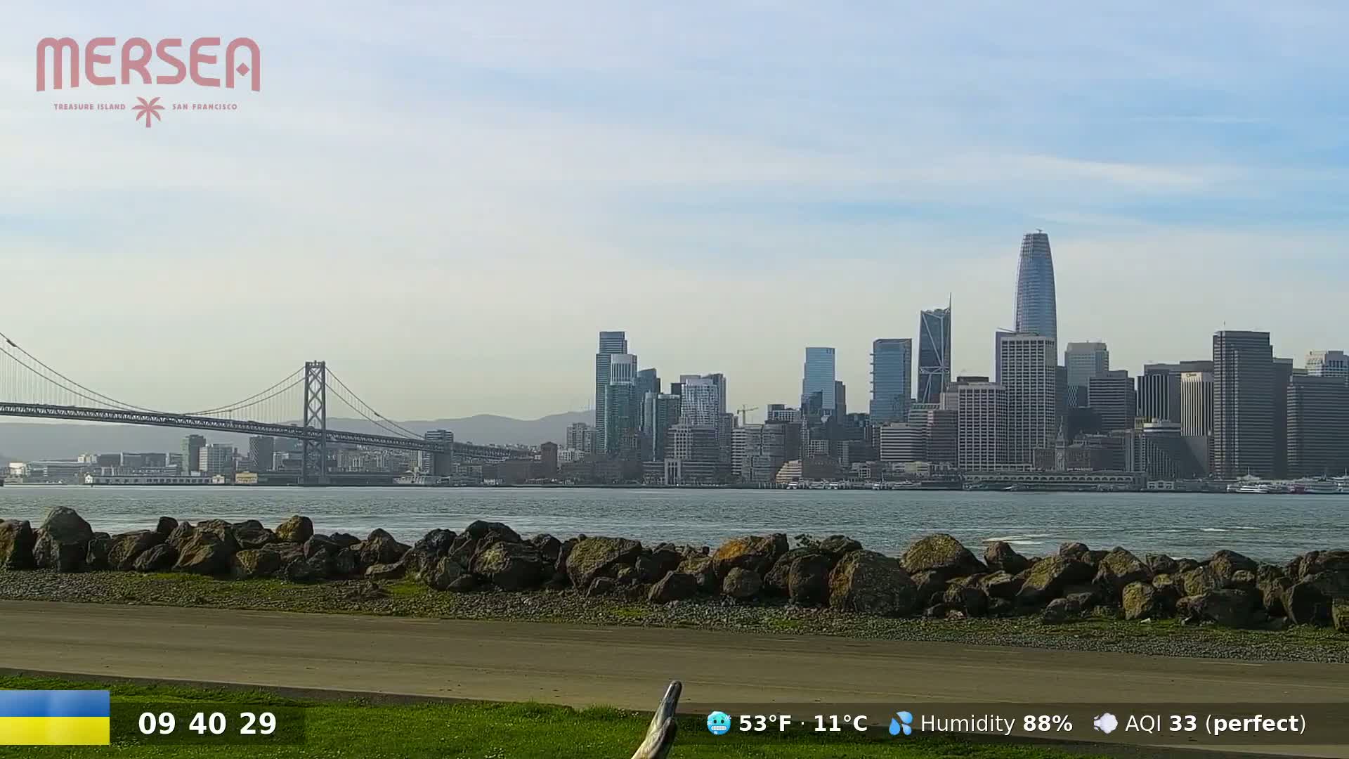 A city skyline with skyscrapers is visible across a body of water from a rocky shoreline under a mostly clear sky.