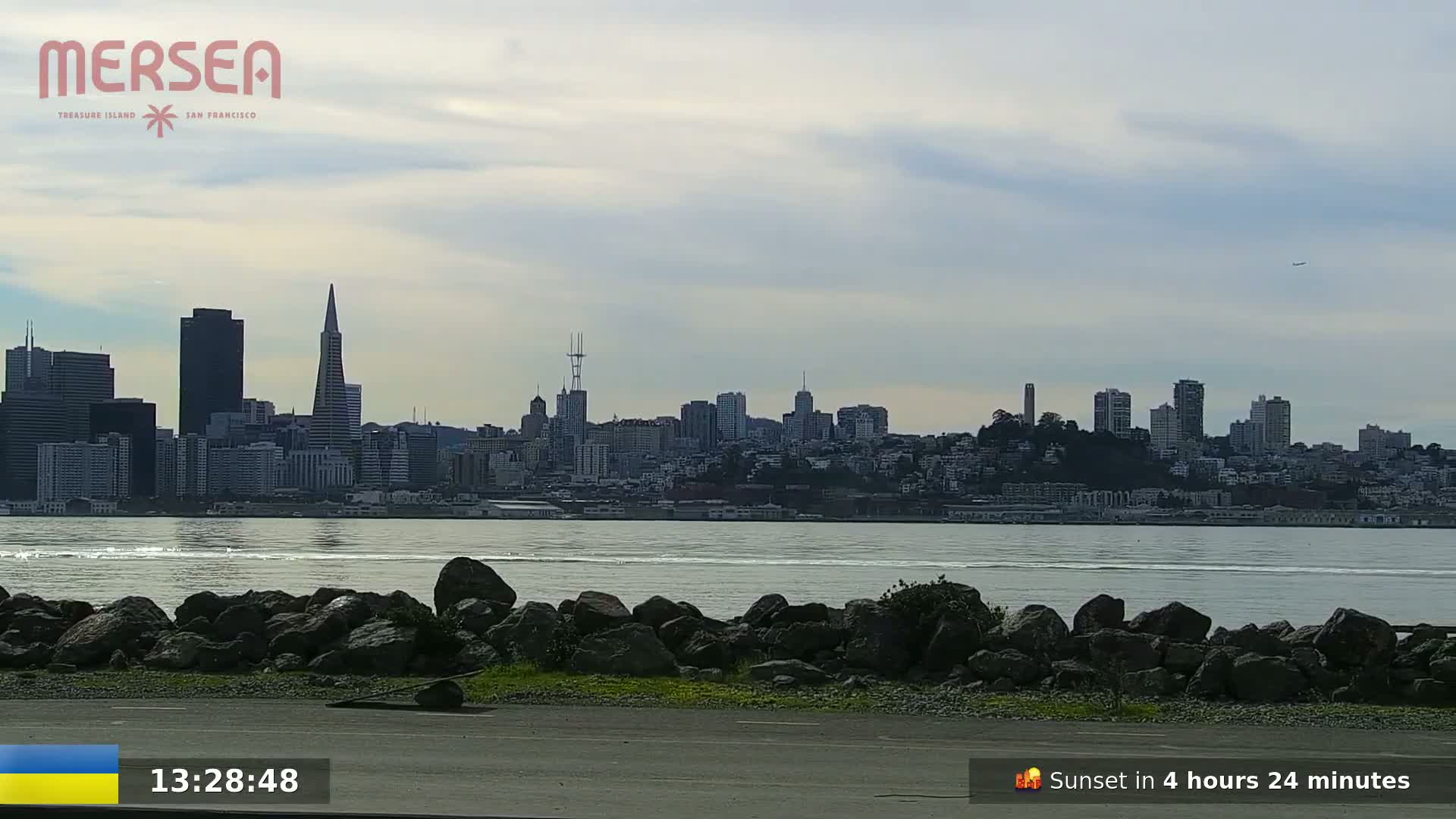 A city skyline is visible across a body of water, under a mostly cloudy sky.