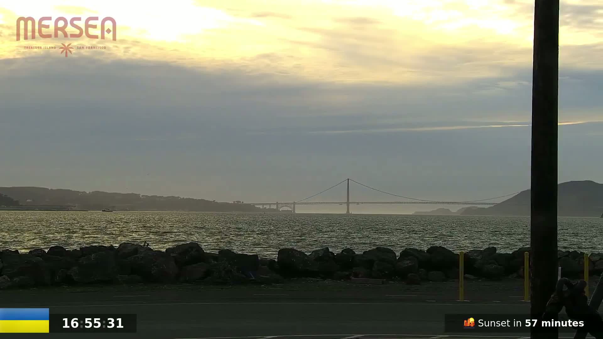 A suspension bridge is visible across a body of water under a partly cloudy, pale yellow sky.