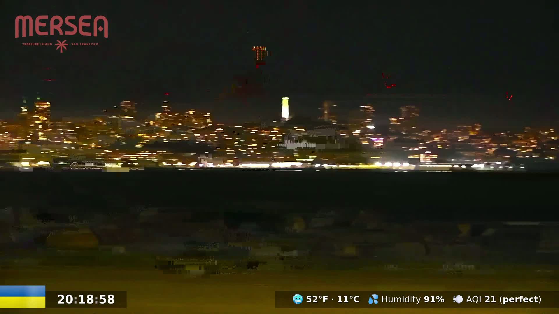 A nighttime view of a city skyline across a body of water, with a dark foreground and high humidity.