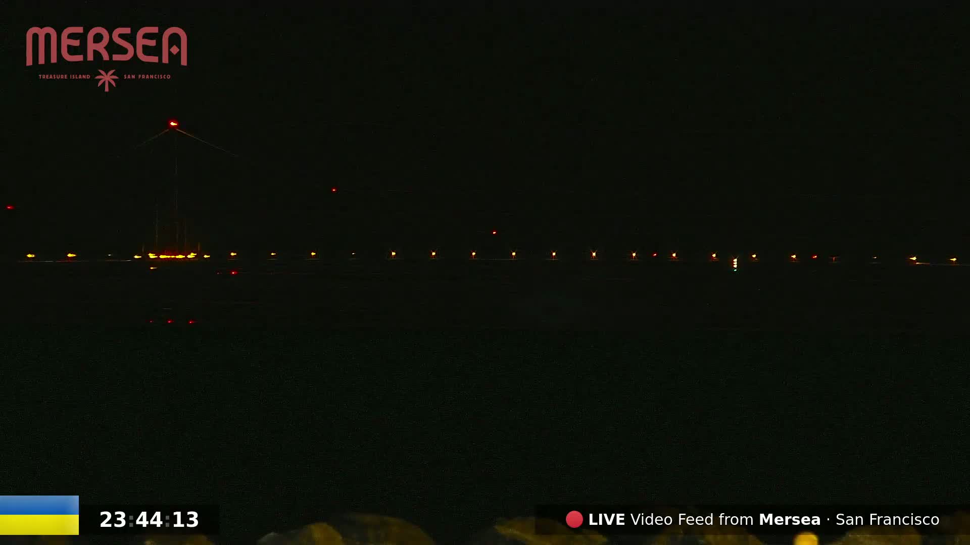 A nighttime long shot shows a dark body of water reflecting a line of lights on a structure in the distance under a clear, dark sky.